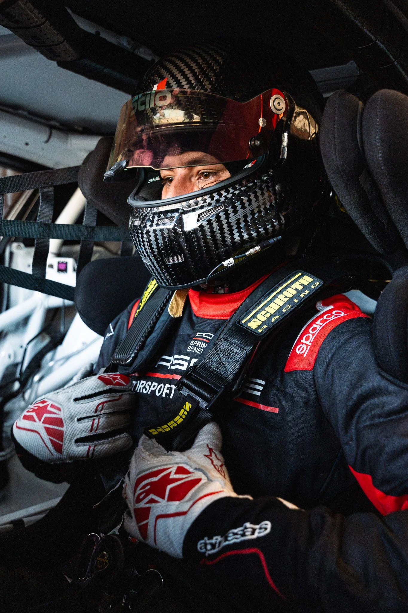 A race car driver in a helmet and racing suit seated inside a race car, preparing for a race.