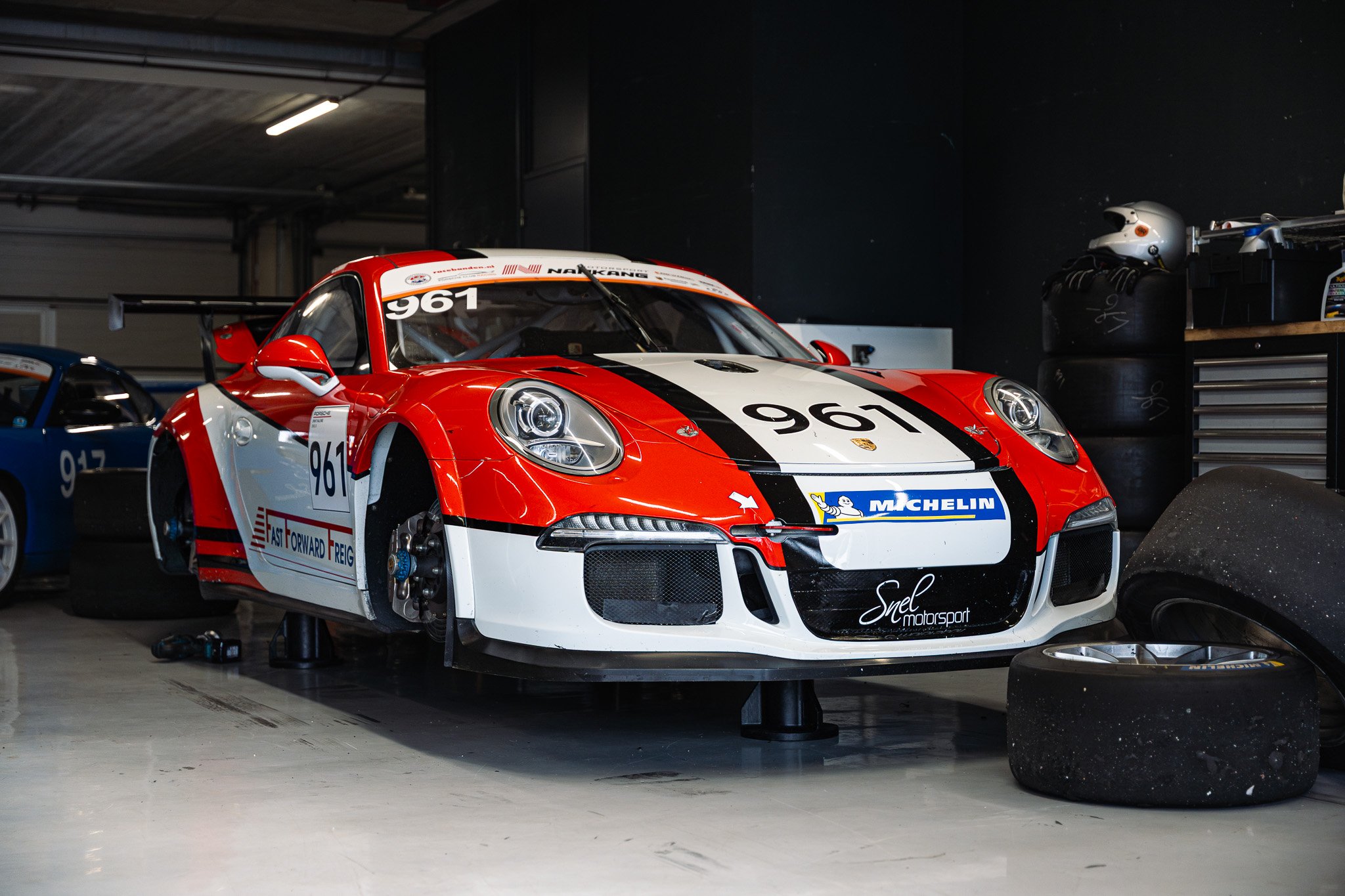 Red and white race car with black stripes, number 961 on the hood and windshield, in a garage with tools and tires.