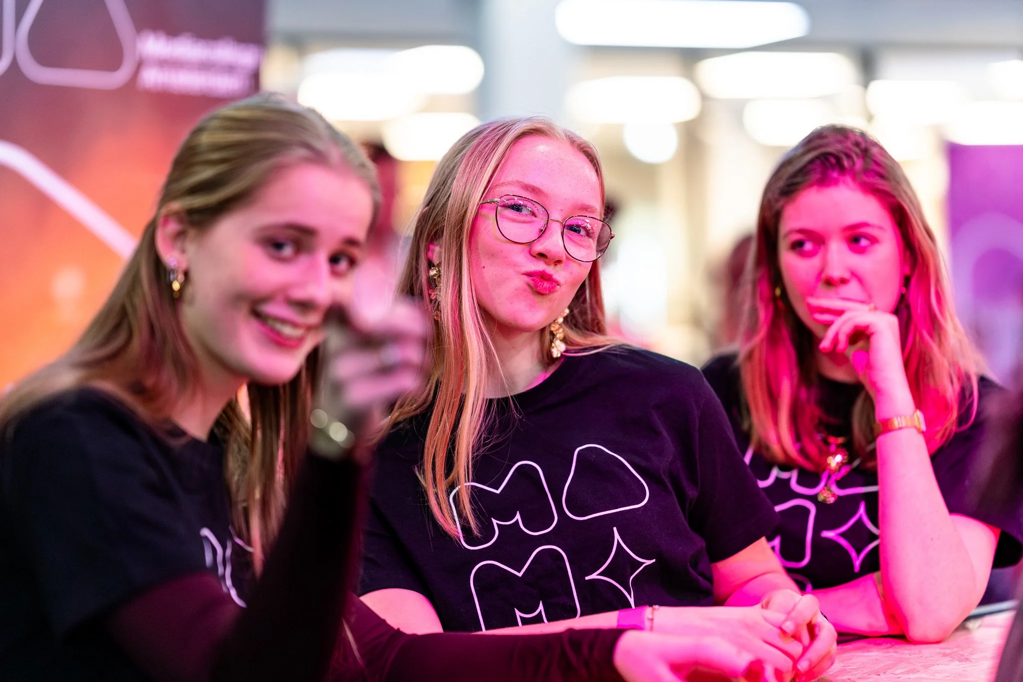 Three young women with long hair sitting at a table, wearing black t-shirts with white designs, in a well-lit indoor setting, some looking at the camera, one making a kissing face, the others with different expressions.