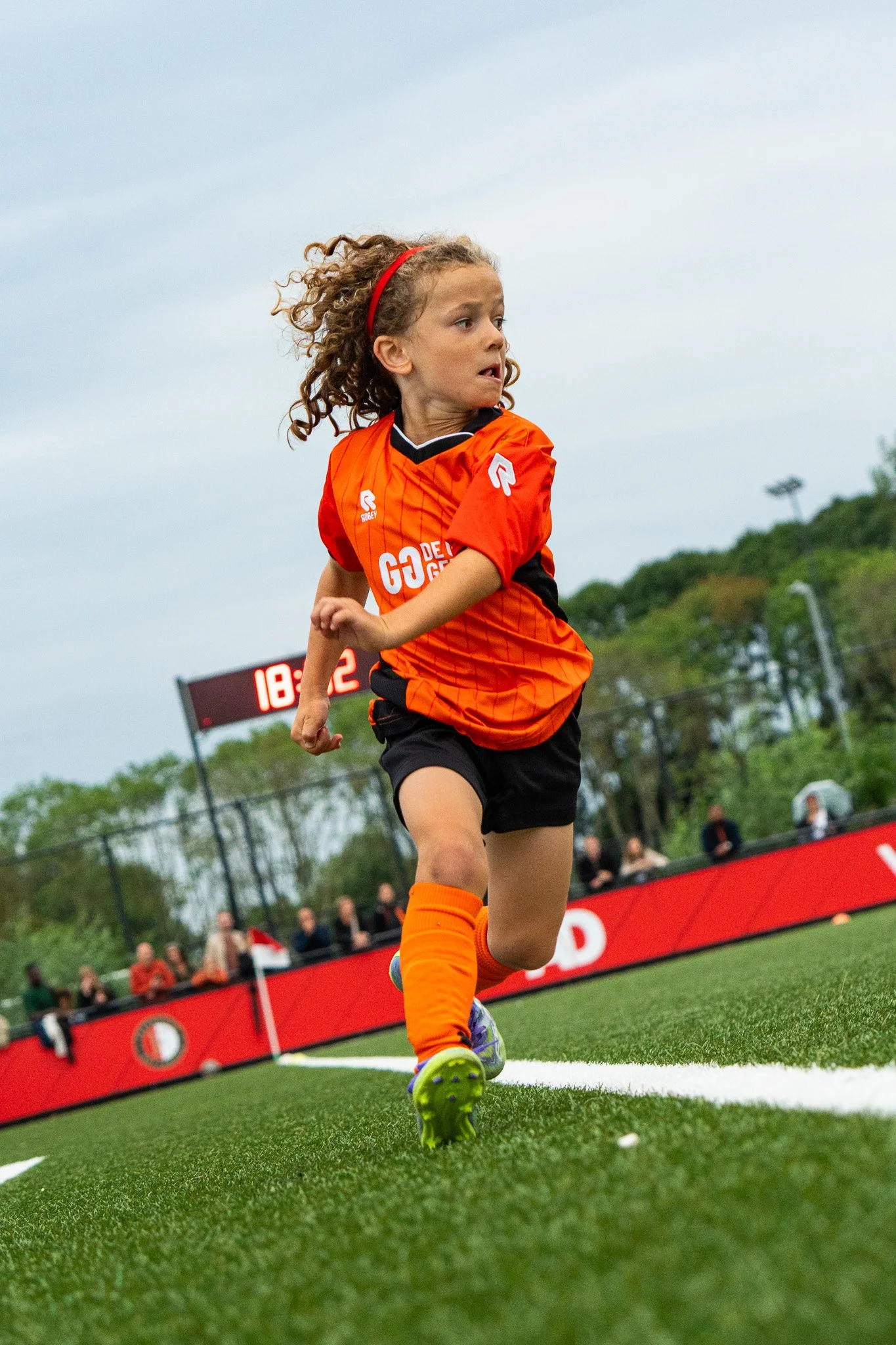 Young girl playing soccer on a field, wearing an orange uniform, running with determination, with a scoreboard and spectators in the background.