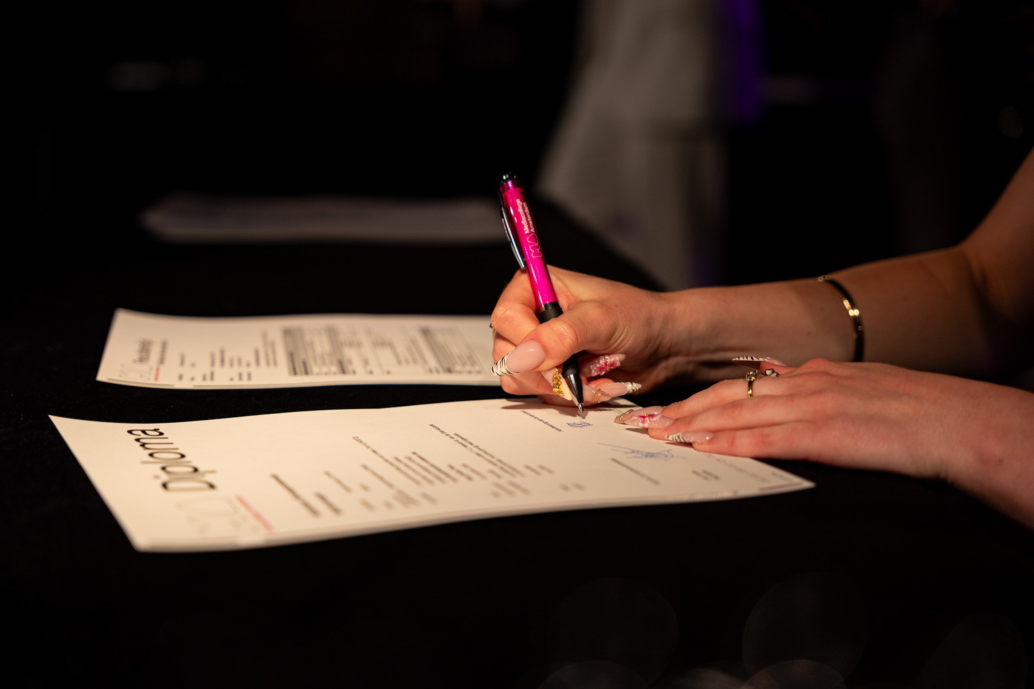 A person signing a document with a pink pen on a black surface.
