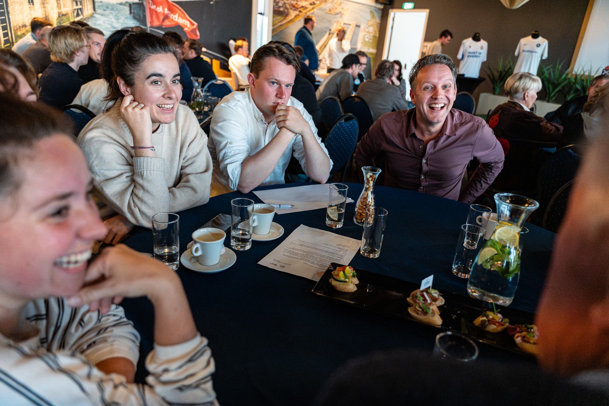 People sitting around a table during an event, smiling and engaging with each other, with drinks and food on the table.