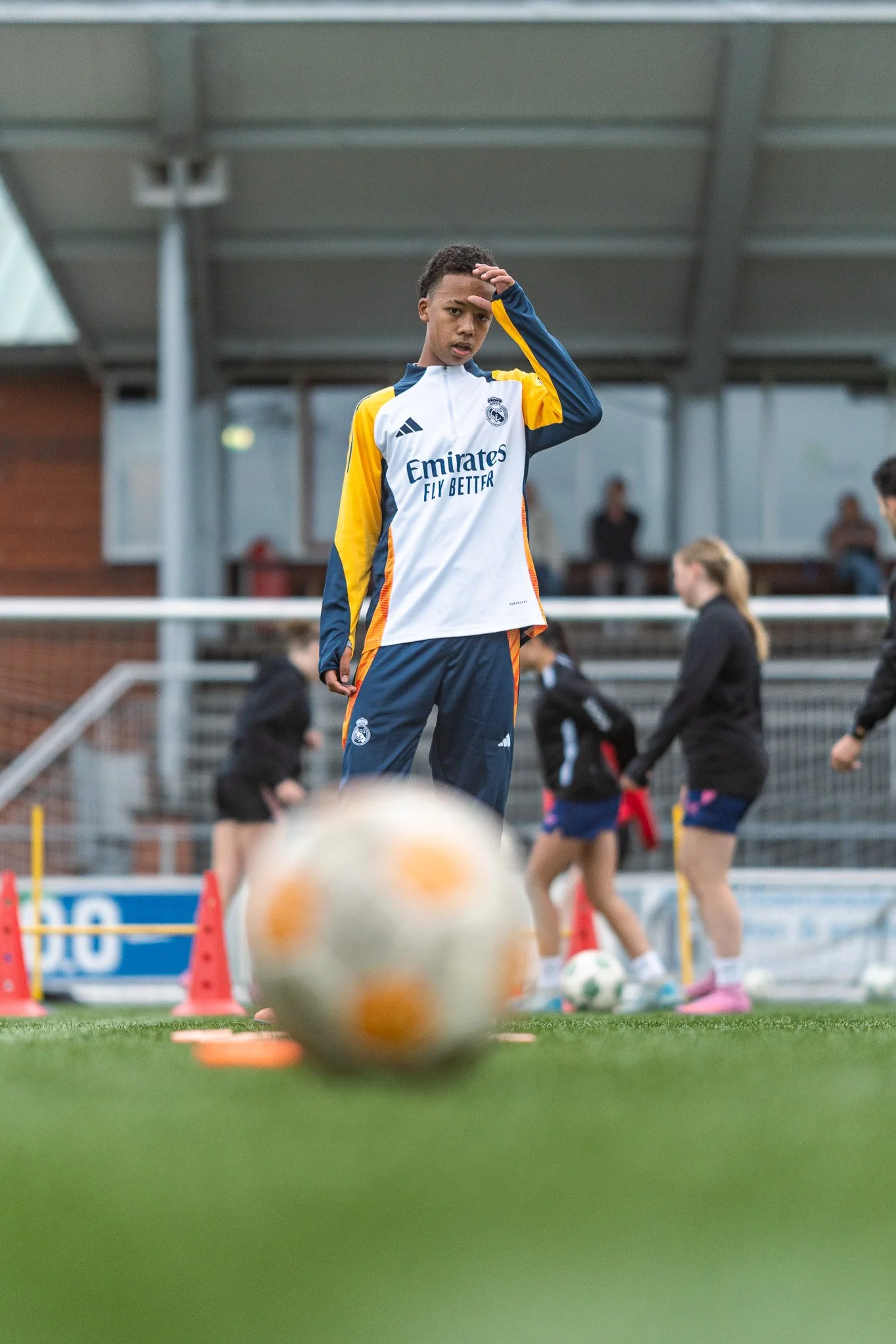 A young male soccer player in a Real Madrid training tracksuit stands on a soccer field, looking at a soccer ball in the foreground, during a practice session with other children in the background.