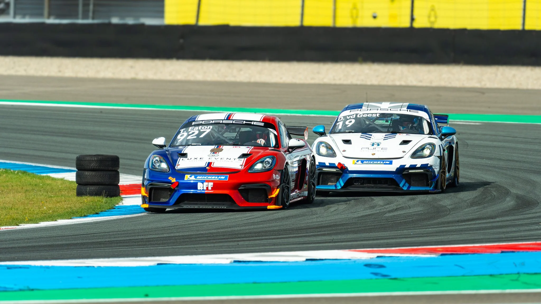 Two Porsche race cars on a race track, making a turn, with one car slightly ahead of the other. The cars are blue, white, and red, with racing numbers 927 and 719, and are surrounded by tire barriers and colorful track markings.
