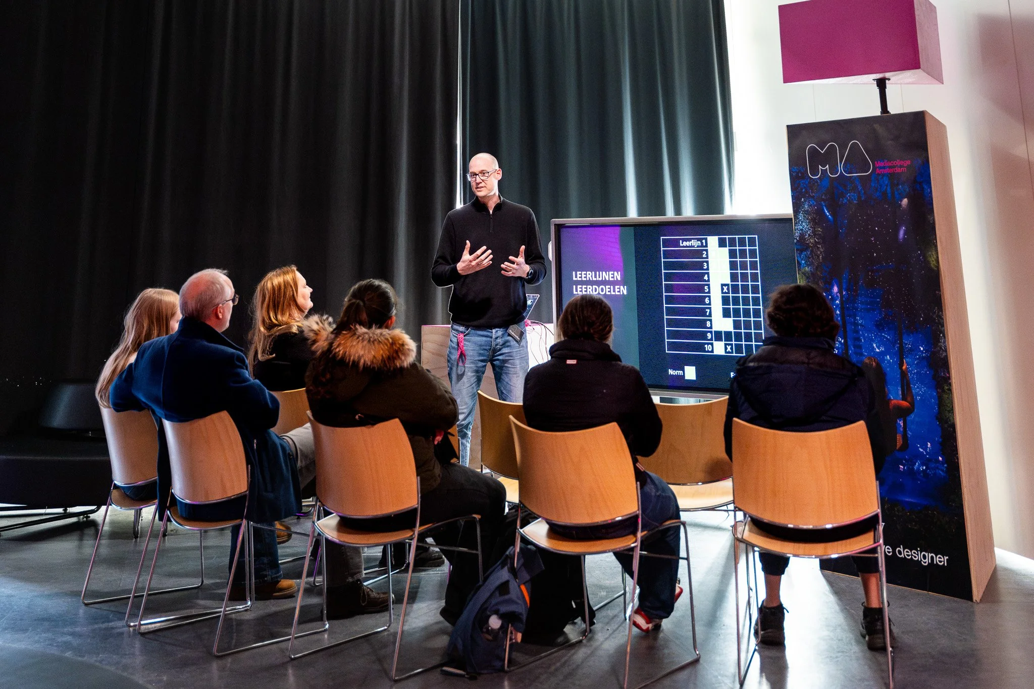 A man giving a presentation to a group of people seated in chairs. The presentation is displayed on a large screen with text in Dutch and grid graphics. The setting appears to be a conference or workshop room with dark curtains and colorful signage.