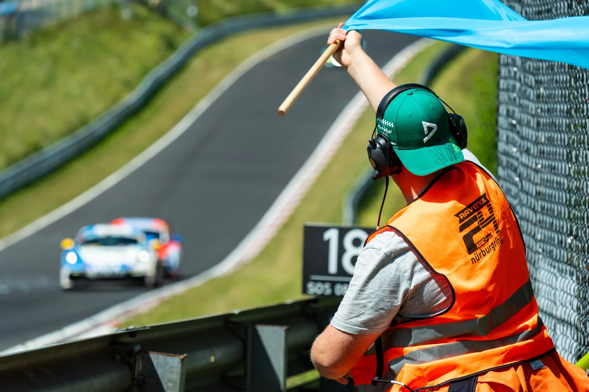 A race official wearing a green cap, orange safety vest, and hearing protection on the side of a race track, signaling with a blue flag as a white race car with blue and red accents passes by.