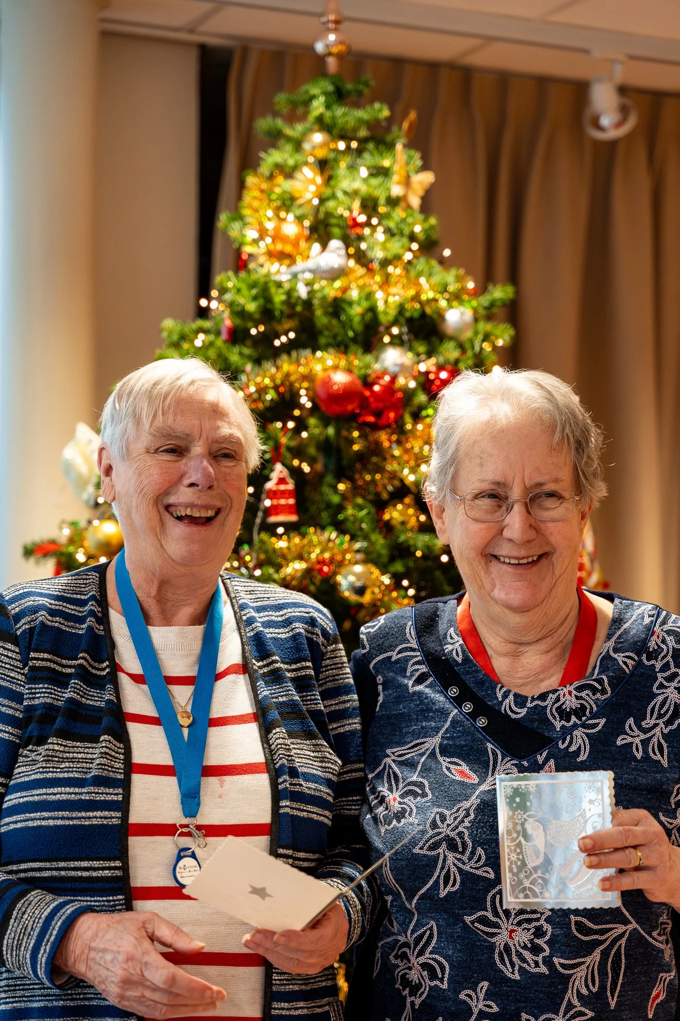 Two elderly women smiling in front of a decorated Christmas tree, holding cards, with festive decorations and ornaments.