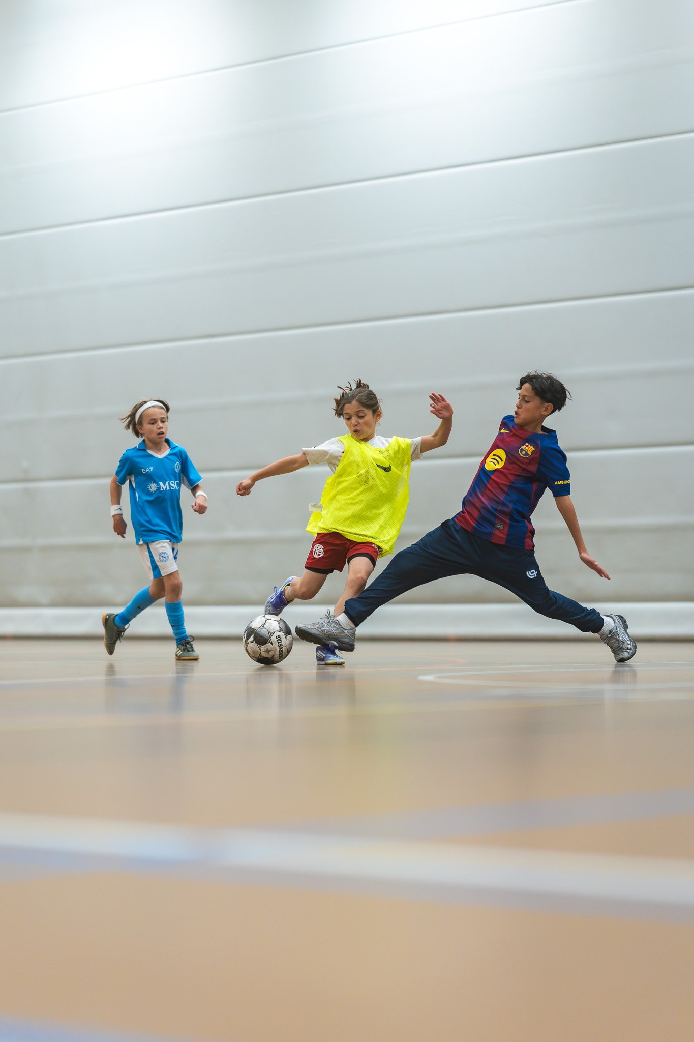 Drie kinderen spelen futsal in een indoor sporthal, een kind in een gele shirt en roodbroek probeert de bal te krijgen van een ander kind in een FC Barcelona shirt, terwijl een ander kind in een blauwe Stella Milenario shirt toekijkt.