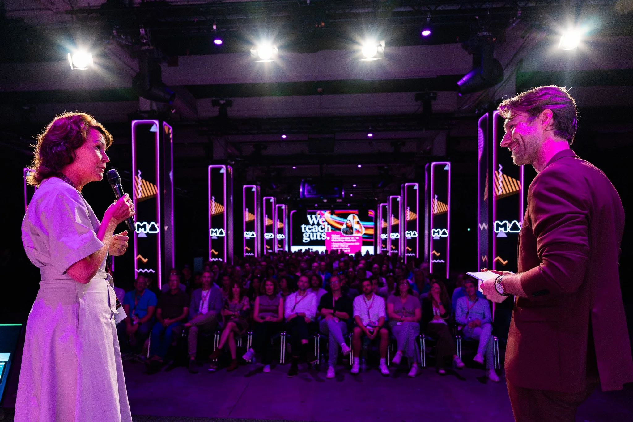 Two speakers, a woman and a man, are speaking on stage at a conference or event, with the woman holding a microphone and the audience seated in front of them. The stage features purple and pink neon lighting.