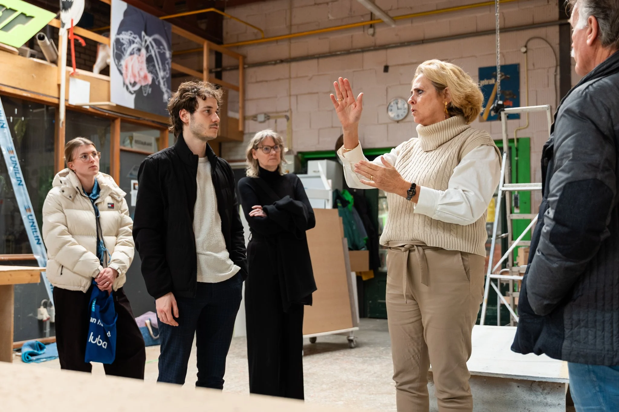 Vijf mensen die naar een vrouw luisteren die iets uitlegt, gelegen in een industriële omgeving.