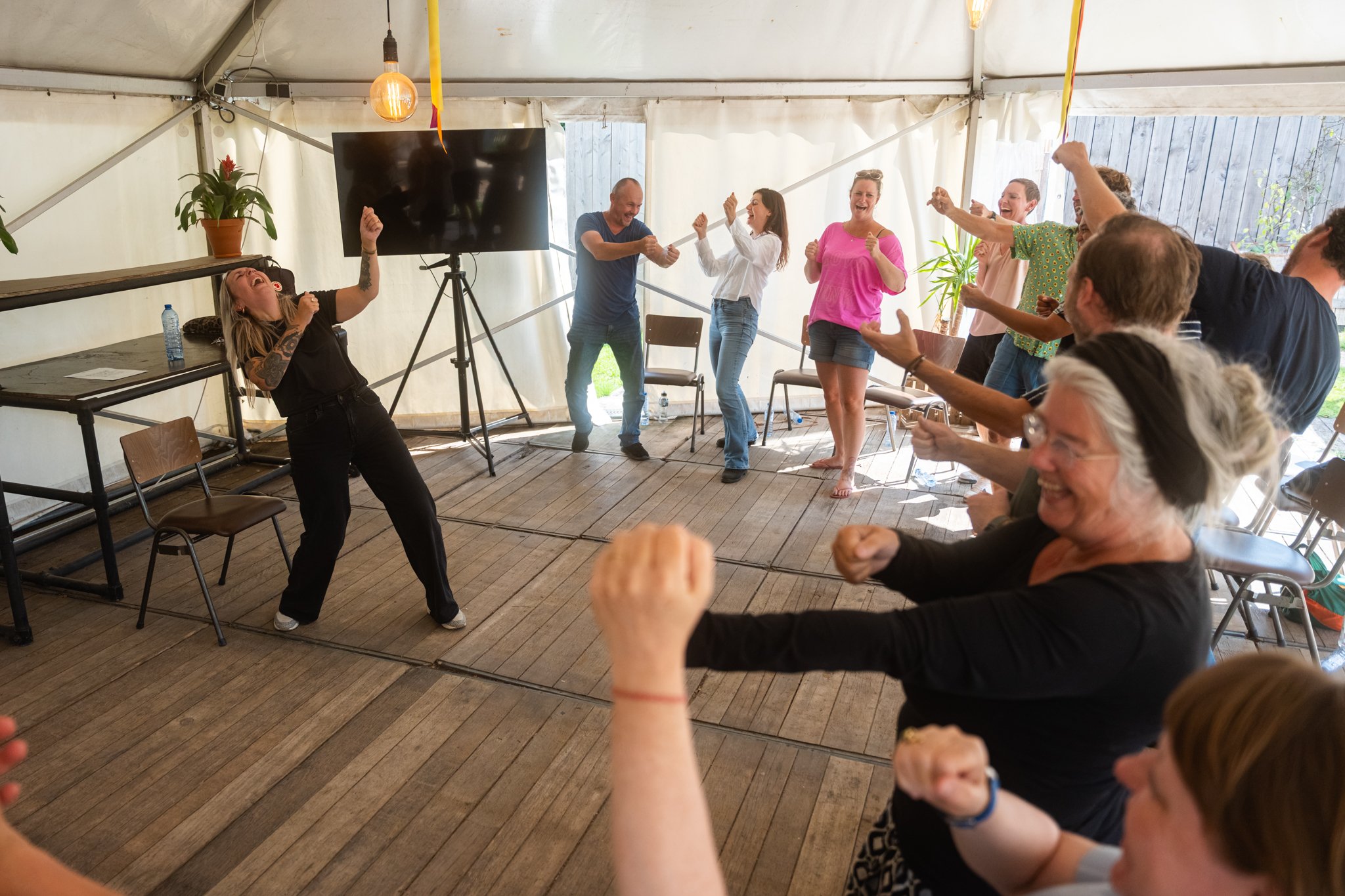 Groep mensen die lachen en dansen bij een feest in een tent, met een vrouw die in het zwart rondloopt en anderen die plezier maken.