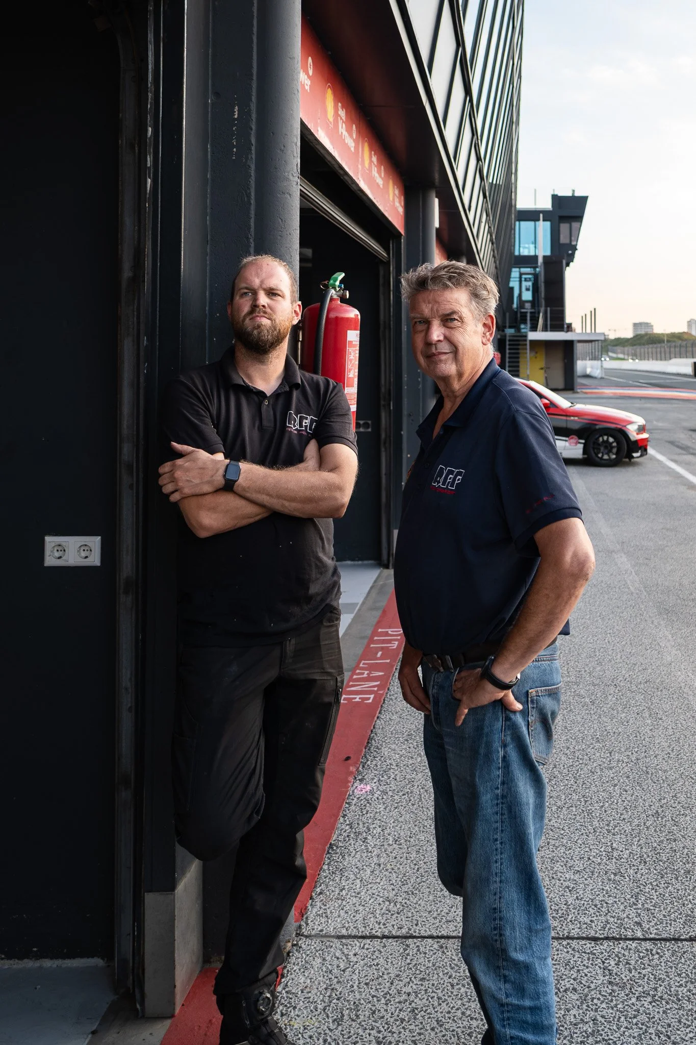 Twee mannen staan naast elkaar buiten op een circuit, een van hen leunt tegen een muur met een brandblusser ernaast, beiden dragen polo's met een logo. Achter hen een raceauto en een modern gebouw.