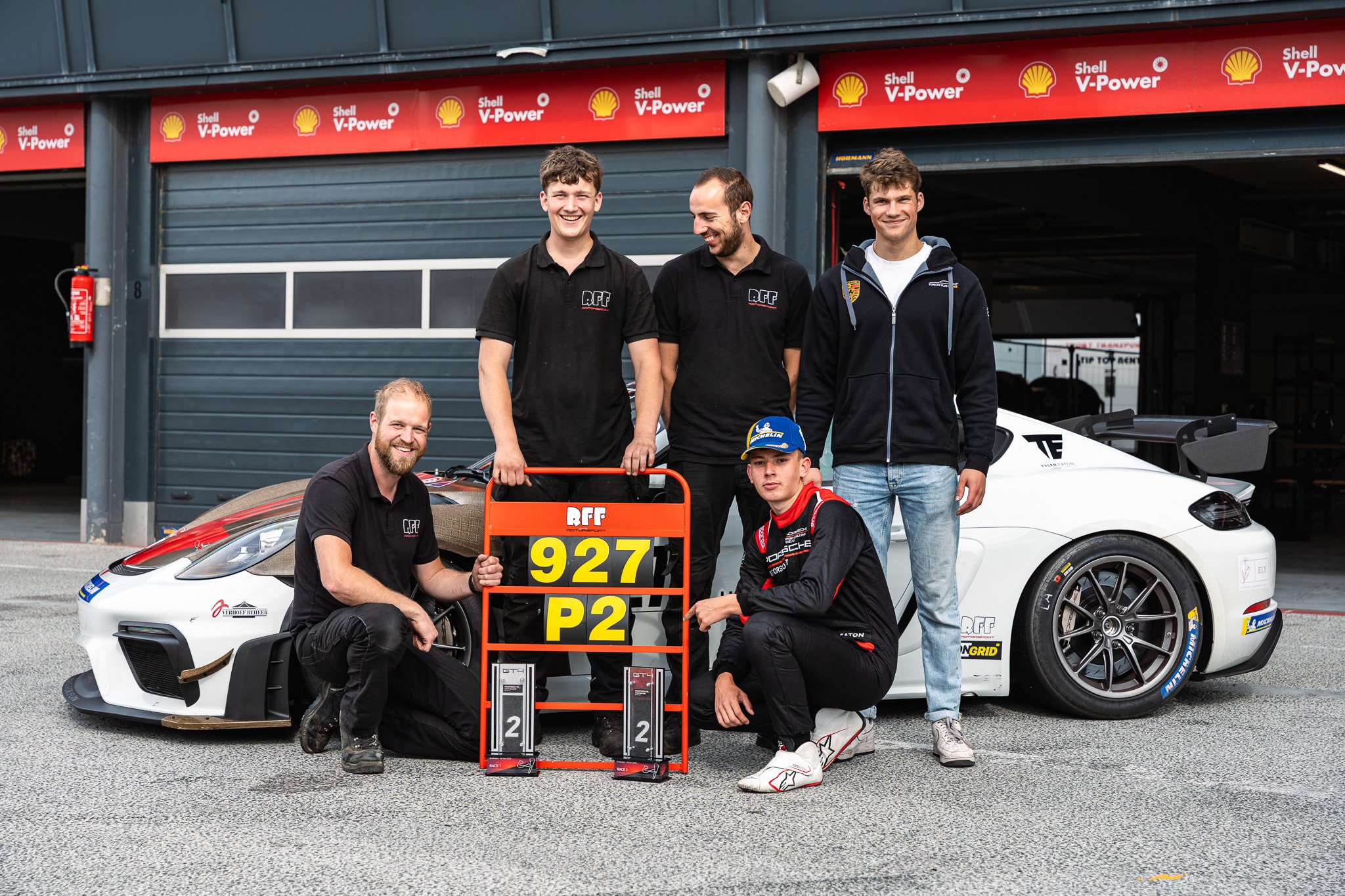 Group of five men in racing gear and team shirts standing and kneeling in front of a white race car at the racing track, with a sign indicating car number 927, class P2, and trophies on the ground.