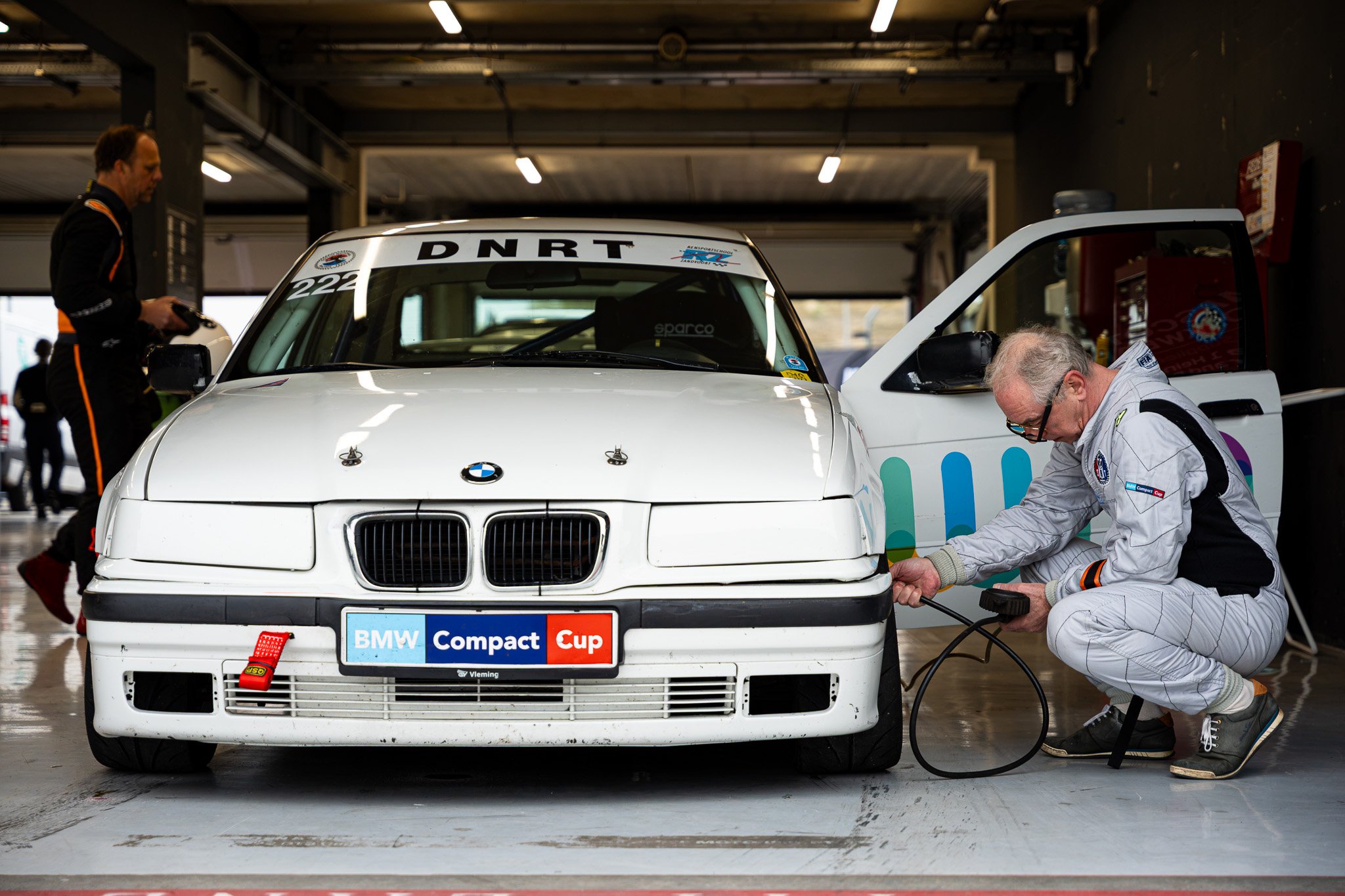 Mensen werken aan een witte BMW raceauto in een garage, met een sticker die zegt 'BMW Compact Cup' op de voorbumper.