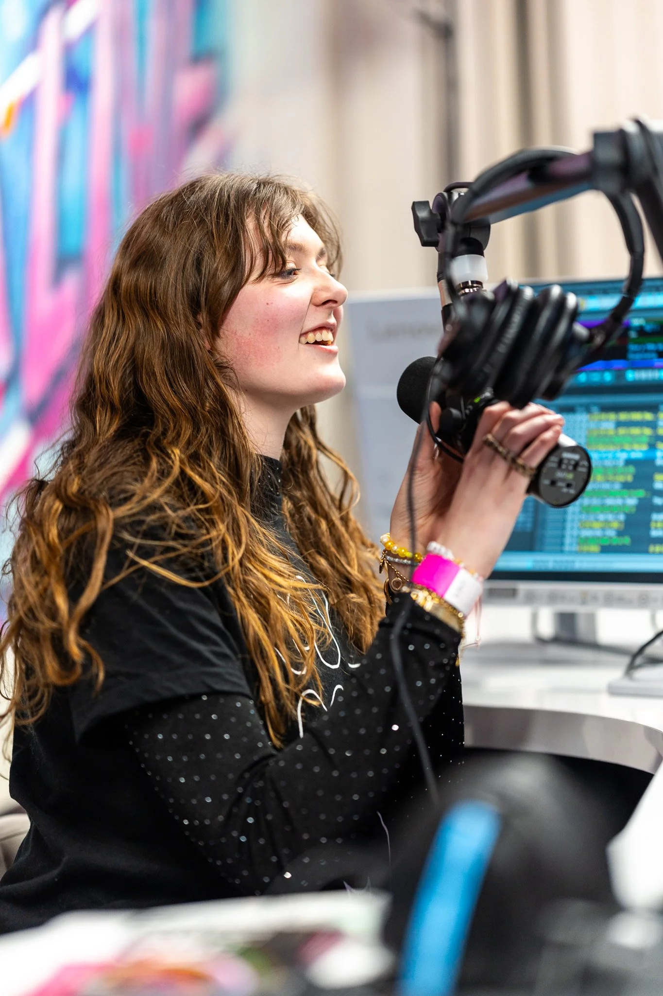 A young woman with long curly red hair holding headphones and microphone in a radio studio, smiling, with a monitor displaying audio waveforms in the background.