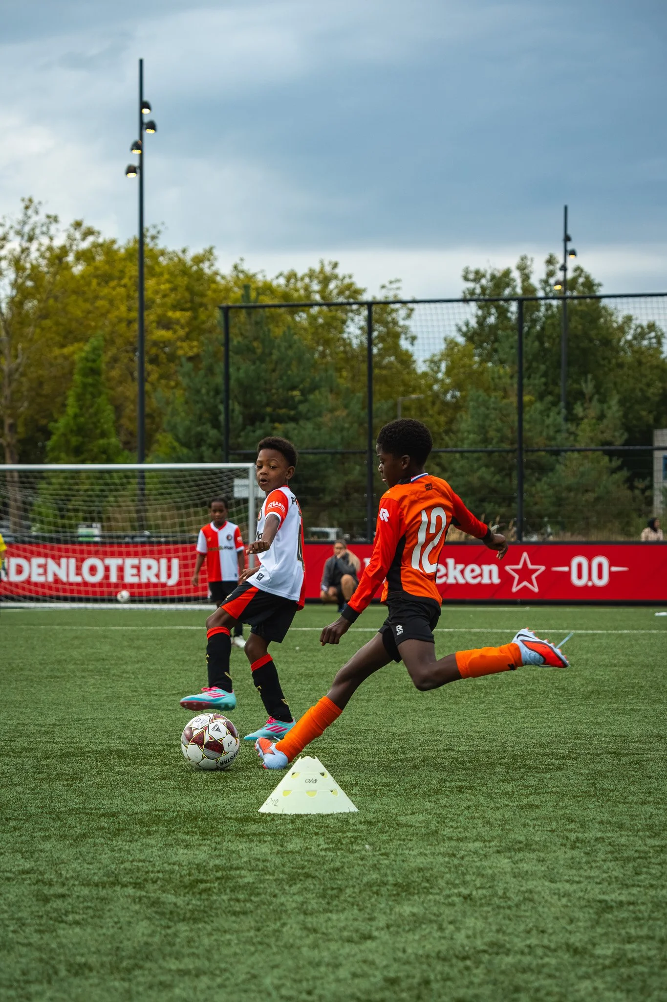 Twee jonge voetballers in een wedstrijd, met een bal op het veld, oefenen sierschot met een witte en gele markeringspilone, terwijl andere kinderen in de achtergrond wachten.