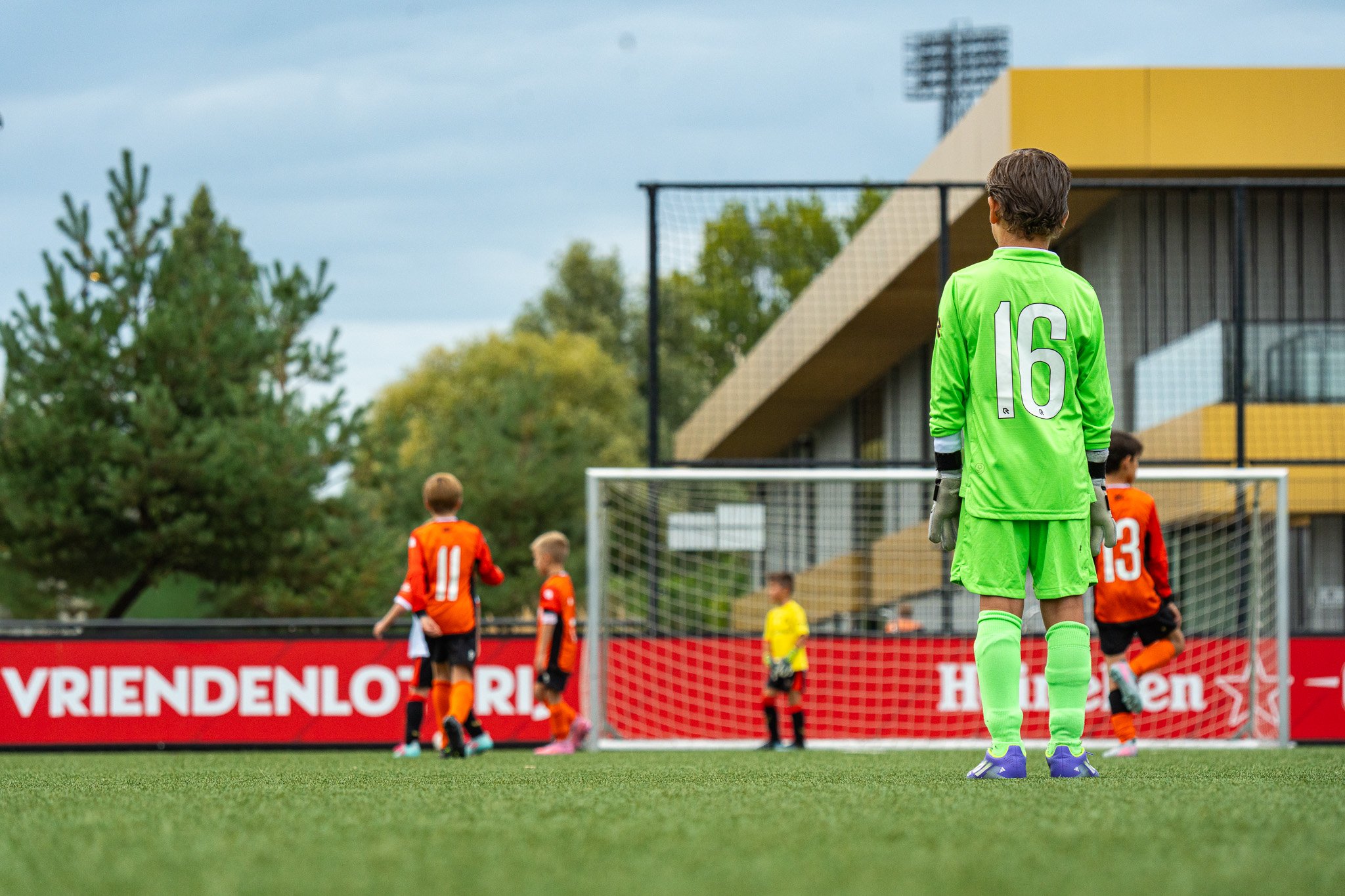 Children playing soccer on a field, with a goalkeeper in a bright green uniform and gloves standing near the goal. Other players are in orange and black uniforms, and a spectator booth is visible in the background.