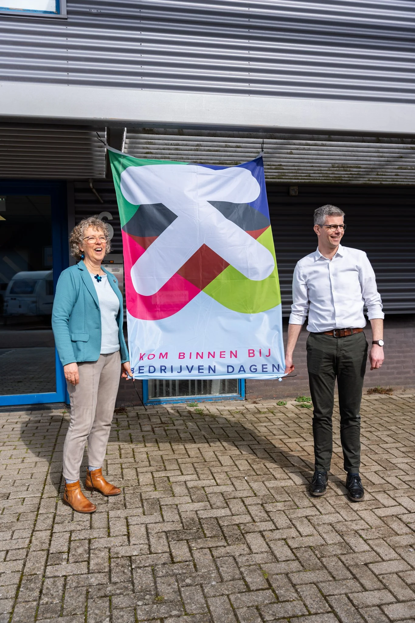 Twee mensen staan buiten, een vrouw en een man, die allebei lachen. Ze houden een banner vast die zegt: 'Kom binnen bij bedrijven dagen'.