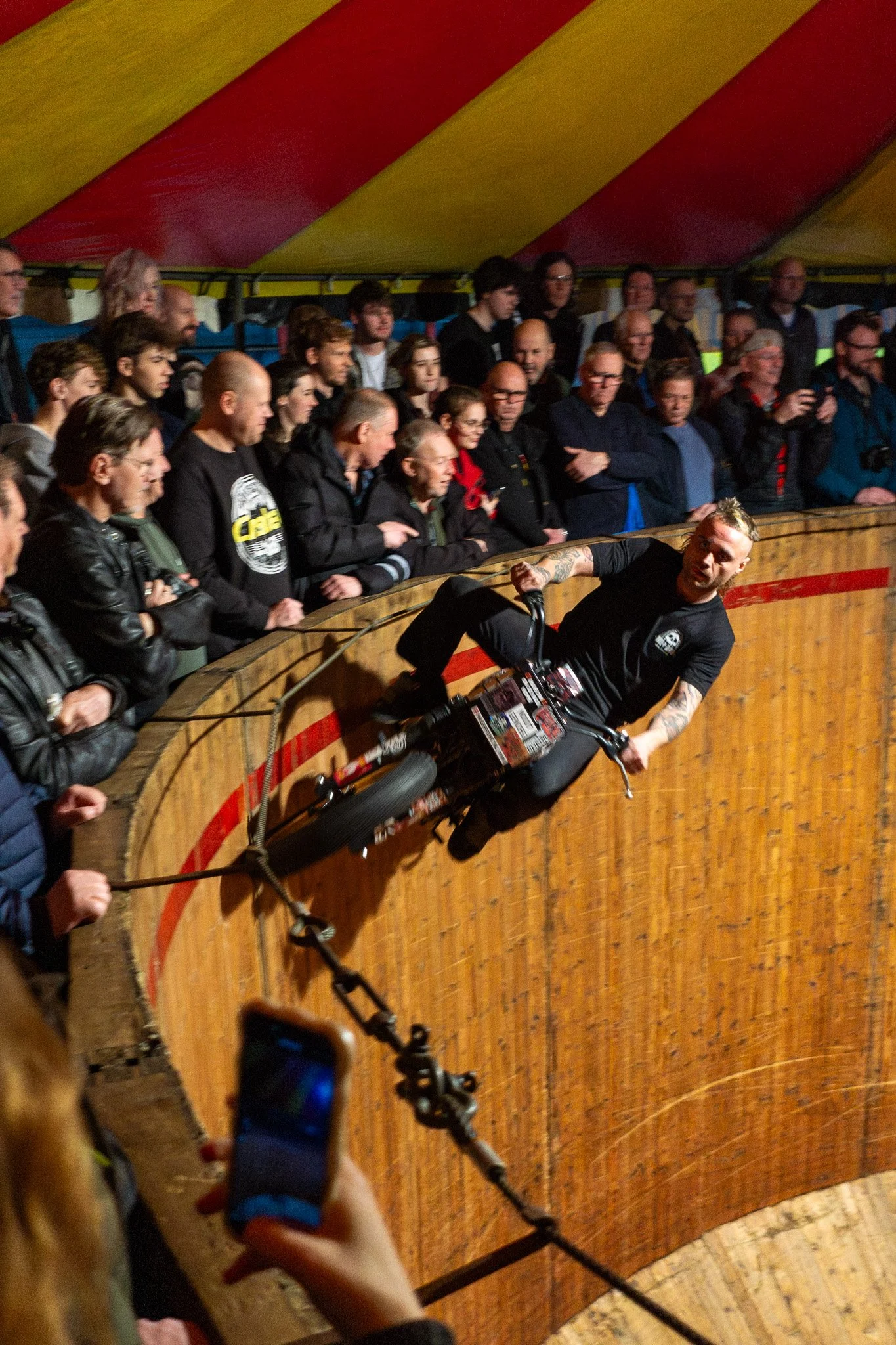 A man riding a BMX bike on a steep wooden curved ramp inside a circus tent, with an audience watching from the side.