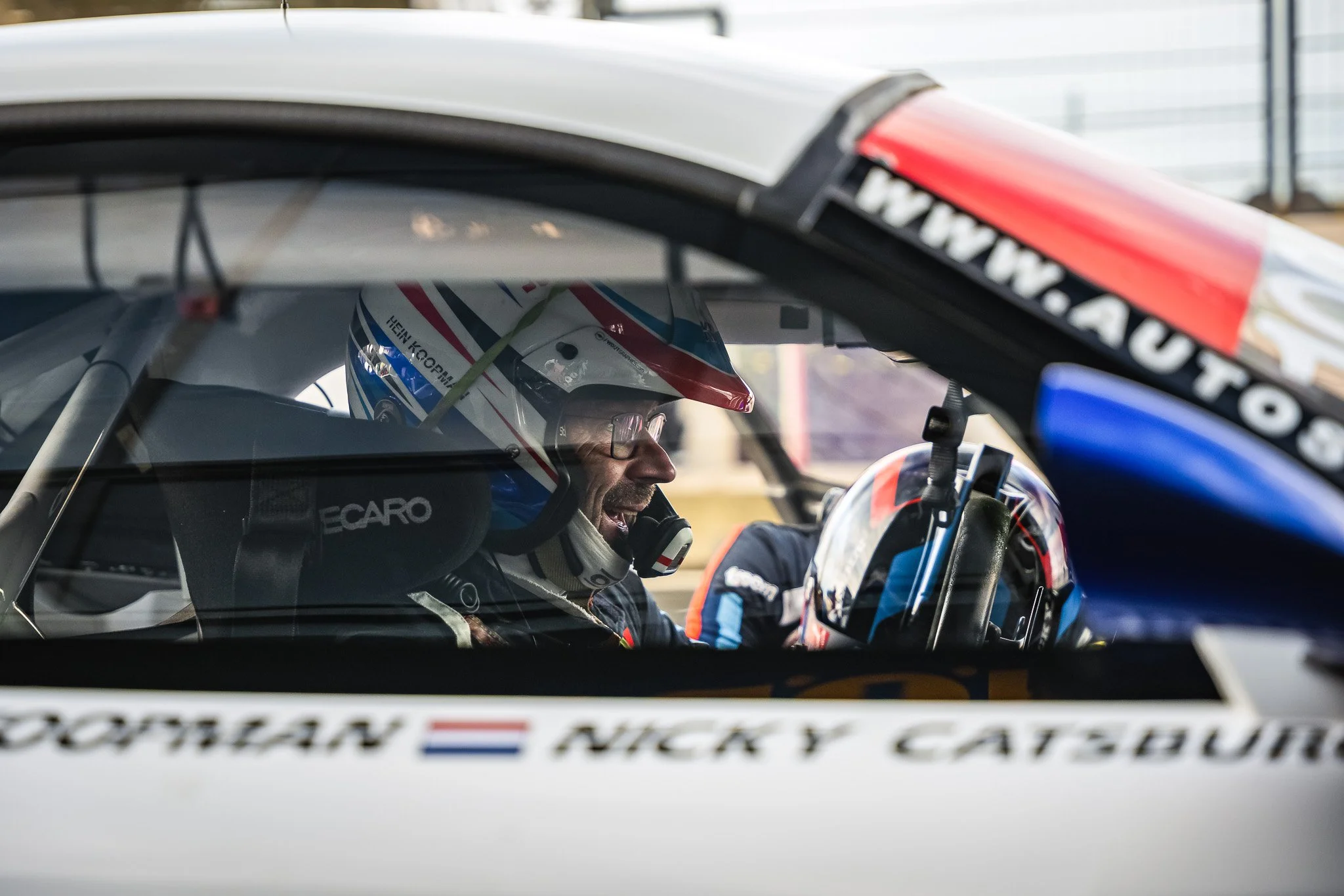 Rennpiloot met helm en oortelefoon in de raceauto, zichtbaar door de raam.