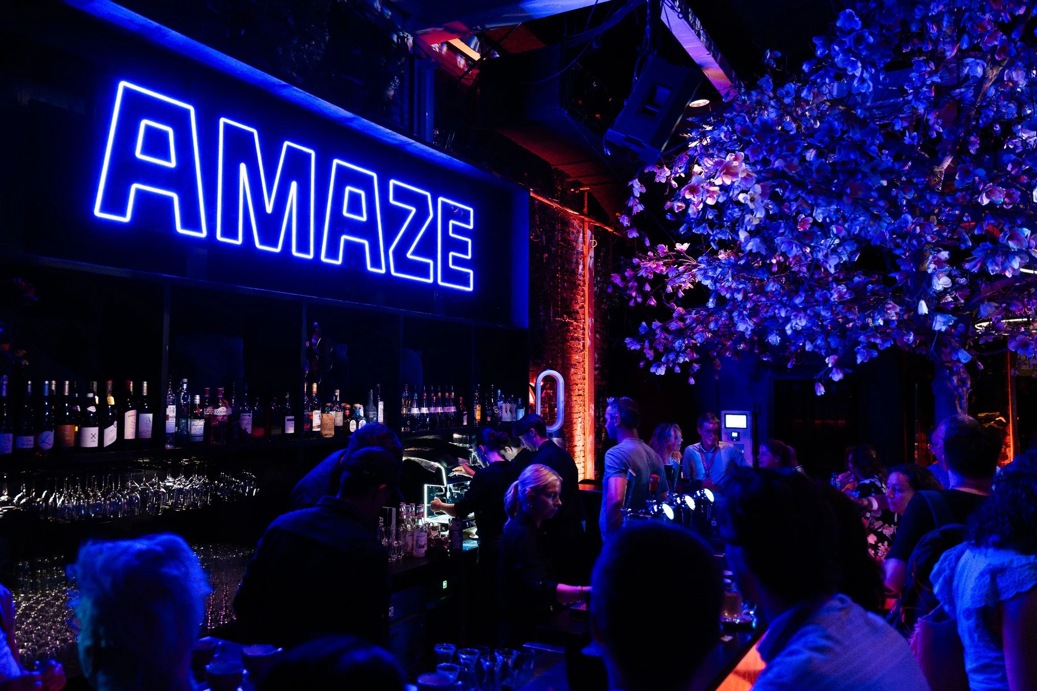 A vibrant bar scene with a large illuminated blue neon sign that reads 'AMAZE,' and a crowd of people socializing, illuminated by colorful neon and purple lighting, with shelves of bottles behind the bar and decorative flowers hanging from the ceilin