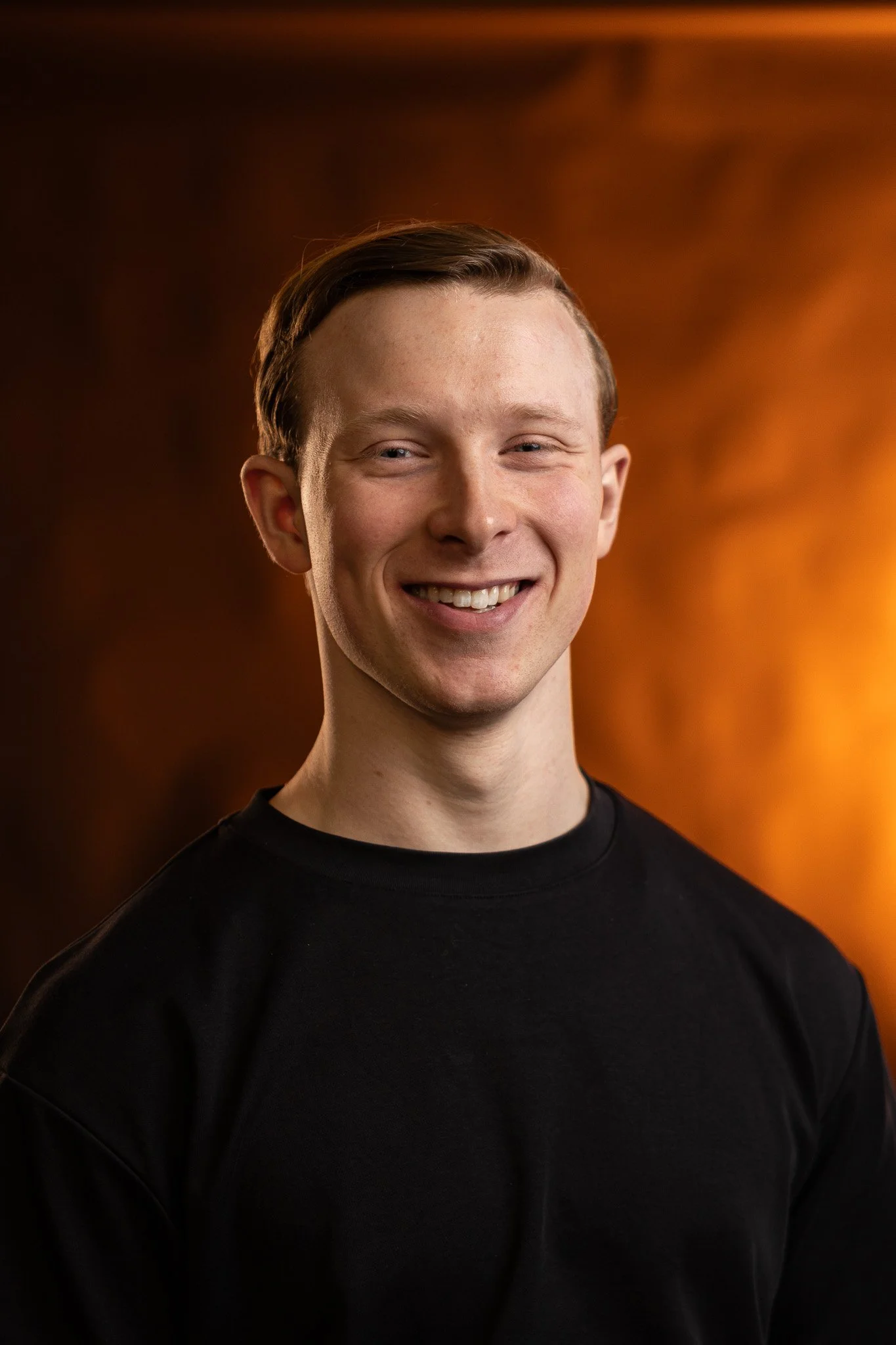 Portret van een jonge man met lichte huidskleur, bruin haar en een brede glimlach, gekleed in een zwarte trui, staand voor een warme, oranje achtergrond.