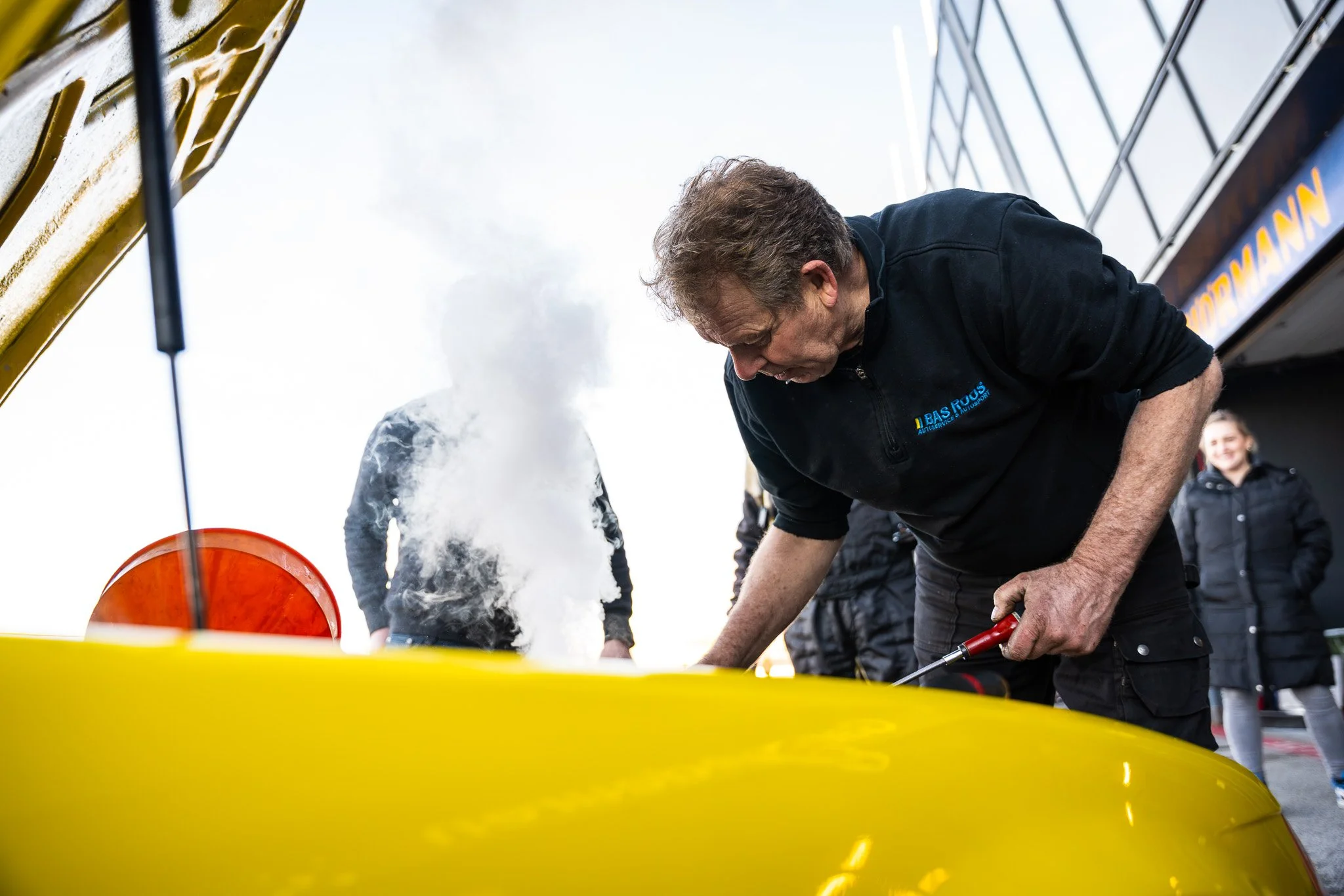 Man in zwart jack die een het maken met een gele raceauto, andere mensen die toekijken, blauwe lucht met wolken.