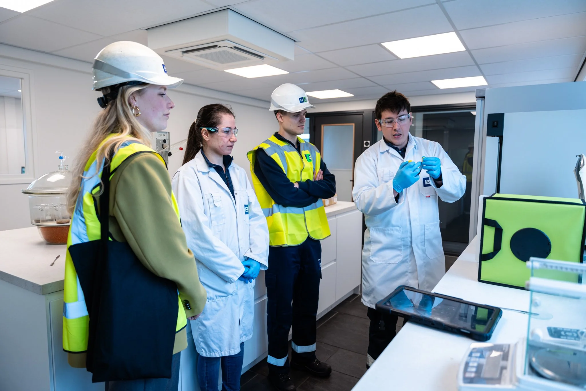 Vier mensen in een laboratorium, drie dragen witte labjassen en veiligheidsbrillen, een draagt een gele veiligheidsjas en een witte helm. Ze kijken naar een onderzoek of demonstratie die door een man met een witte labjas en blauwe handschoenen wordt 