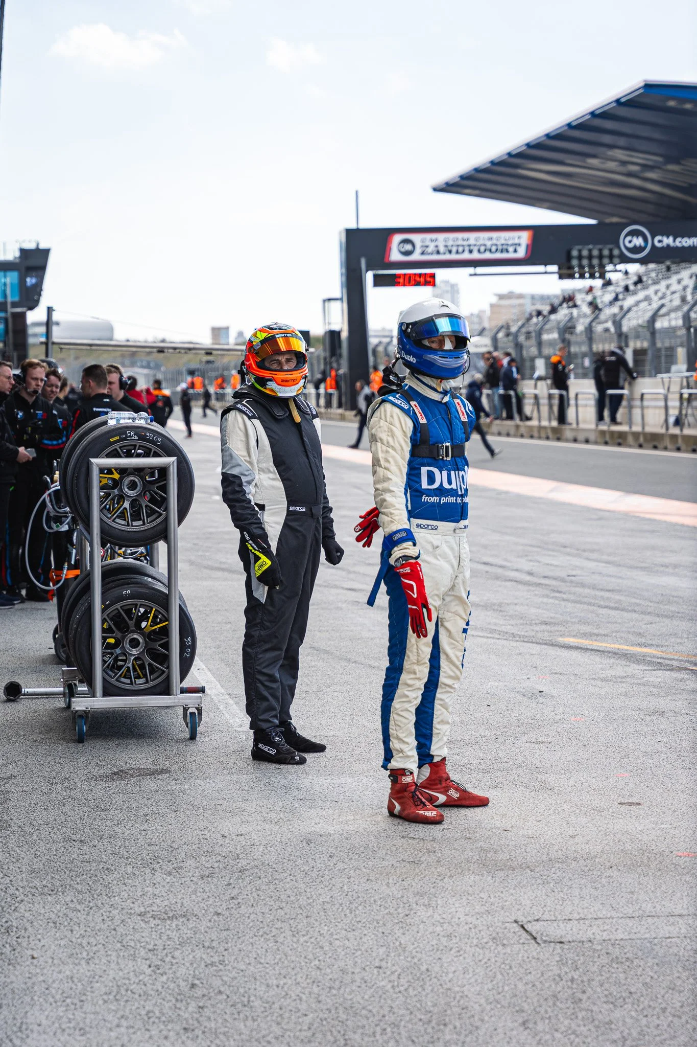 Twee raceauto coureurs in racepakke, klaar om te racen op het circuit. Een coureur draagt een blauwe raceoverall en een ander een zwart-witte raceoverall. Er zijn mensen op de achtergrond en banden die klaarstaan voor gebruik.