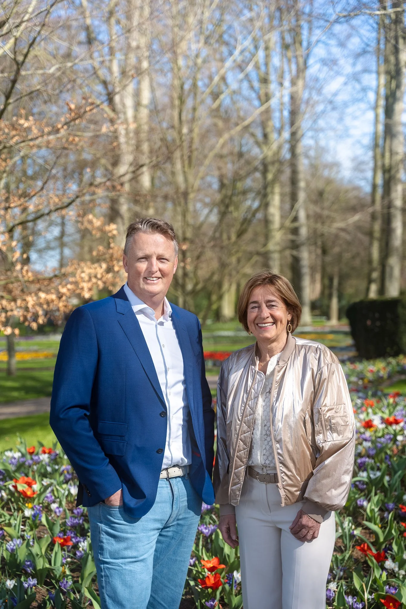 A man and woman standing outdoors in a park with blooming tulips and trees in the background.