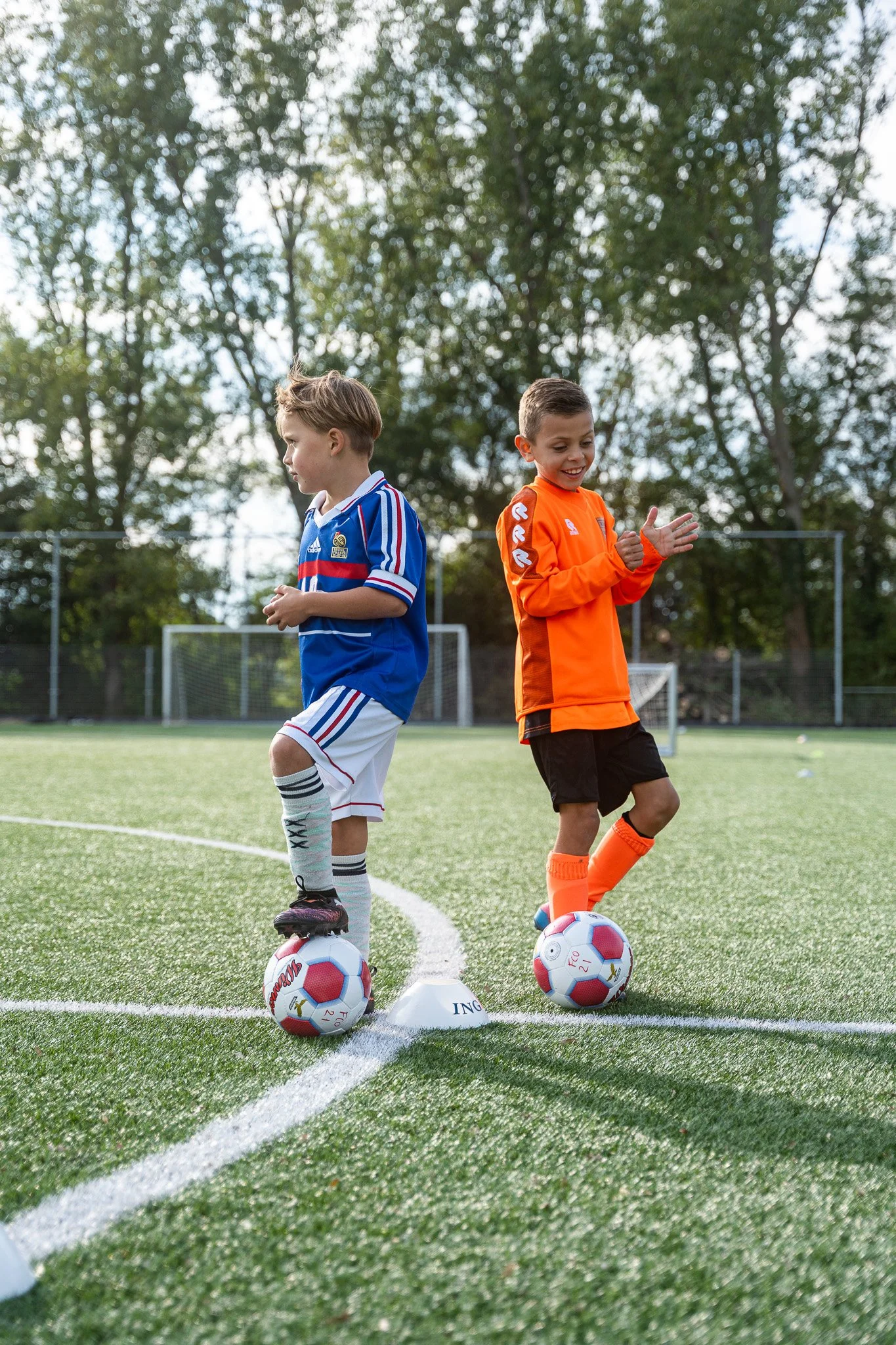 Two young boys standing on a soccer field with soccer balls under their feet, practicing dribbling drills with cones, surrounded by trees.