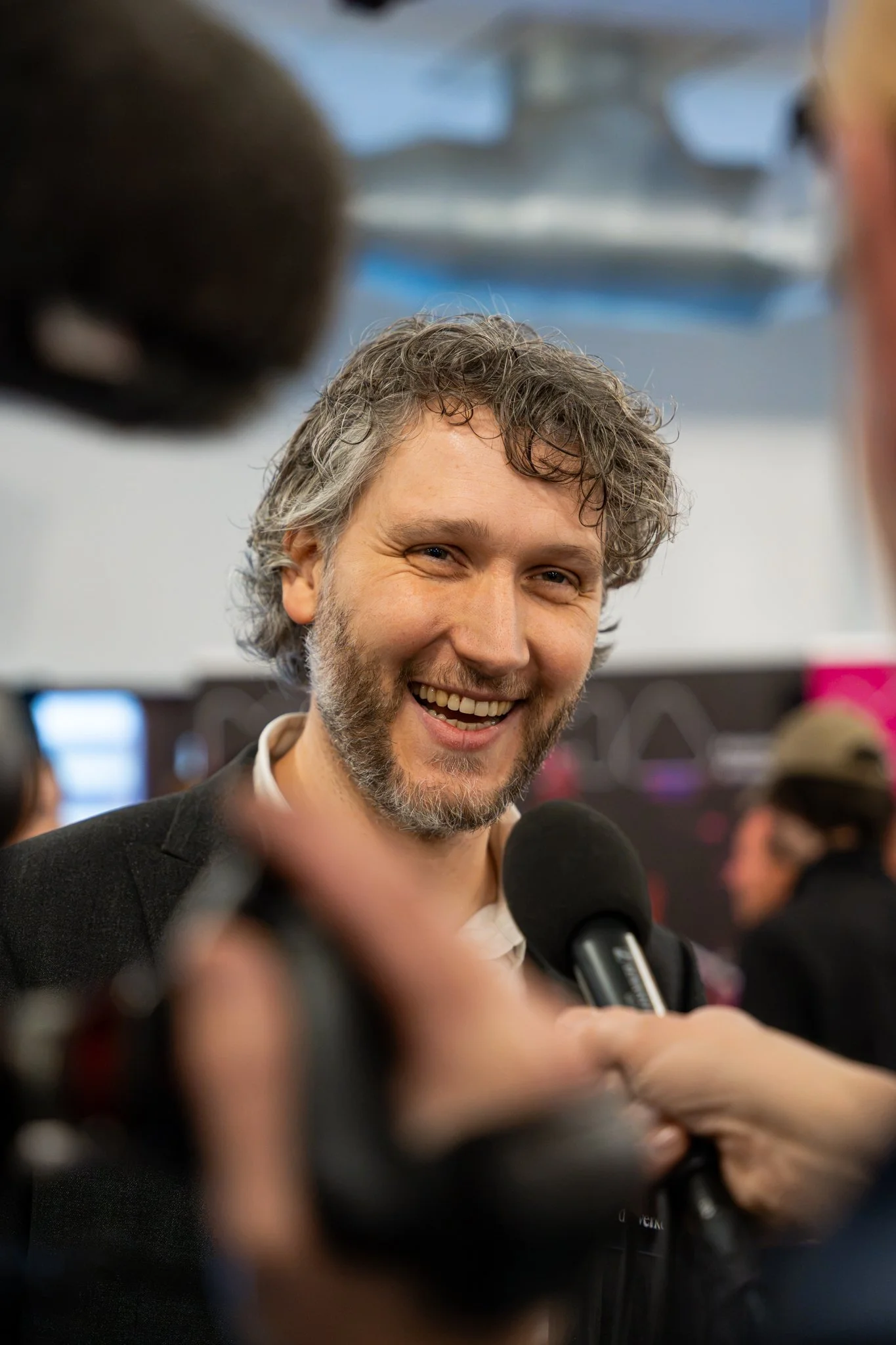 Een mannelijk persoon met grijs, golvend haar en een brede glimlach, omringd door microfoons, tijdens een interview of persconferentie.