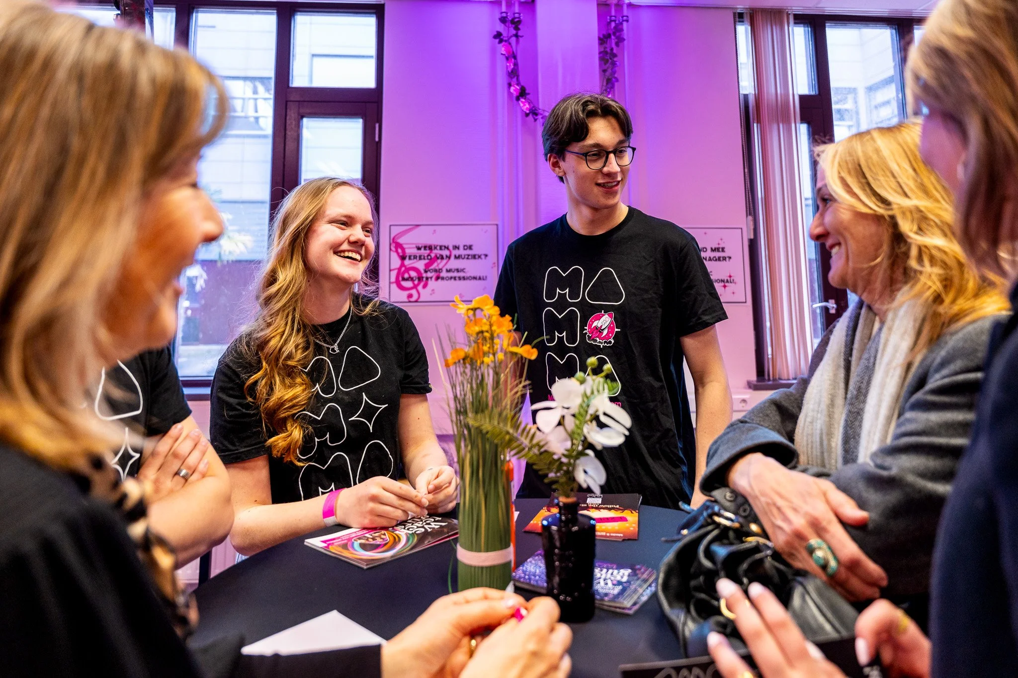Group of young people and adults smiling and talking at a table with flowers in vases, in a room with large windows and purple lighting, during an event.
