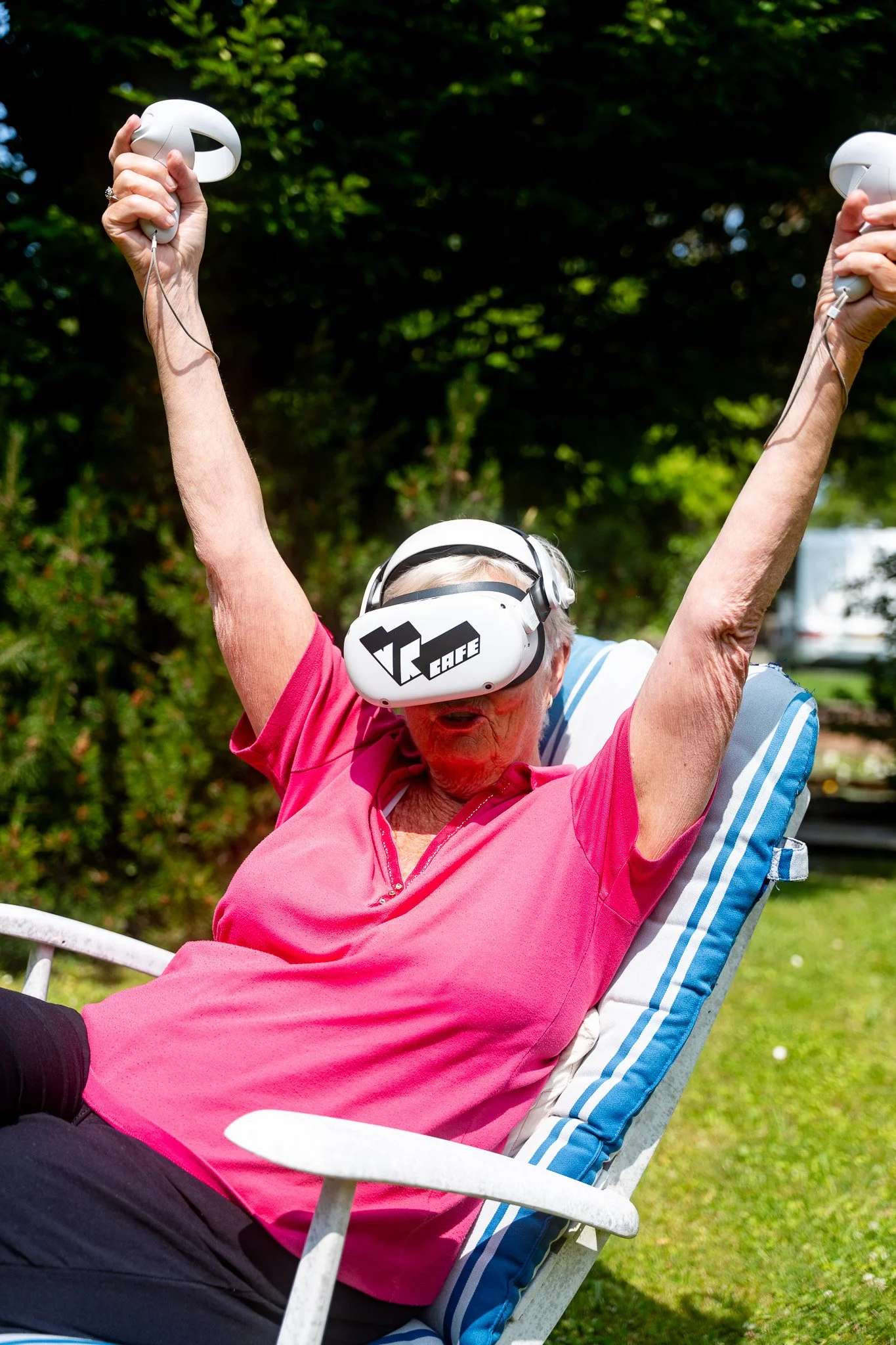 An elderly woman wearing a pink shirt and a VR headset, sitting on a lounge chair outdoors with her arms raised in excitement.