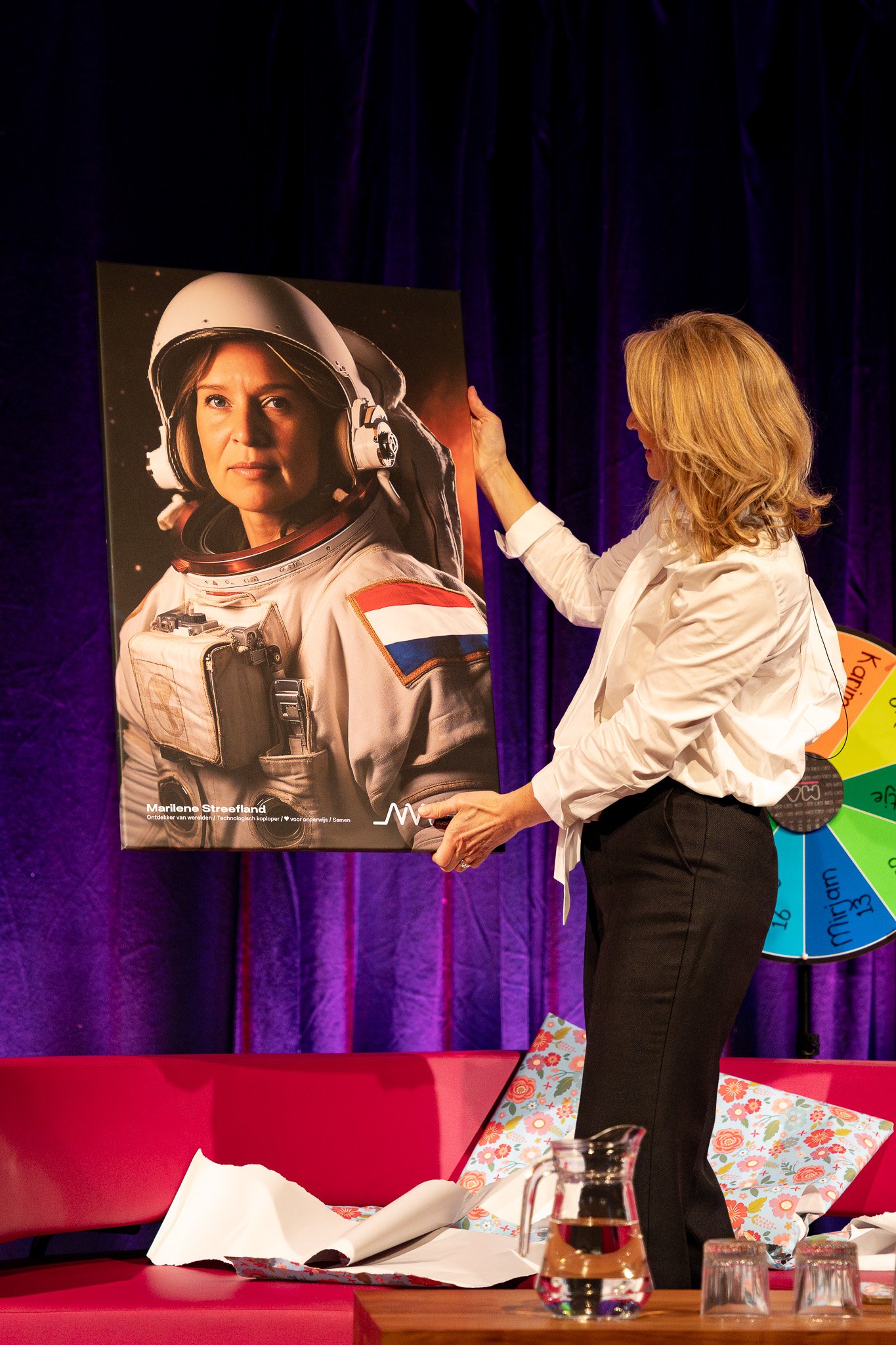 A woman with blonde hair in a white shirt and black pants is holding a large portrait of a female astronaut in a space suit. The background includes a purple curtain, a colorful spinning wheel, pillows on a red couch, and a glass pitcher of water on 