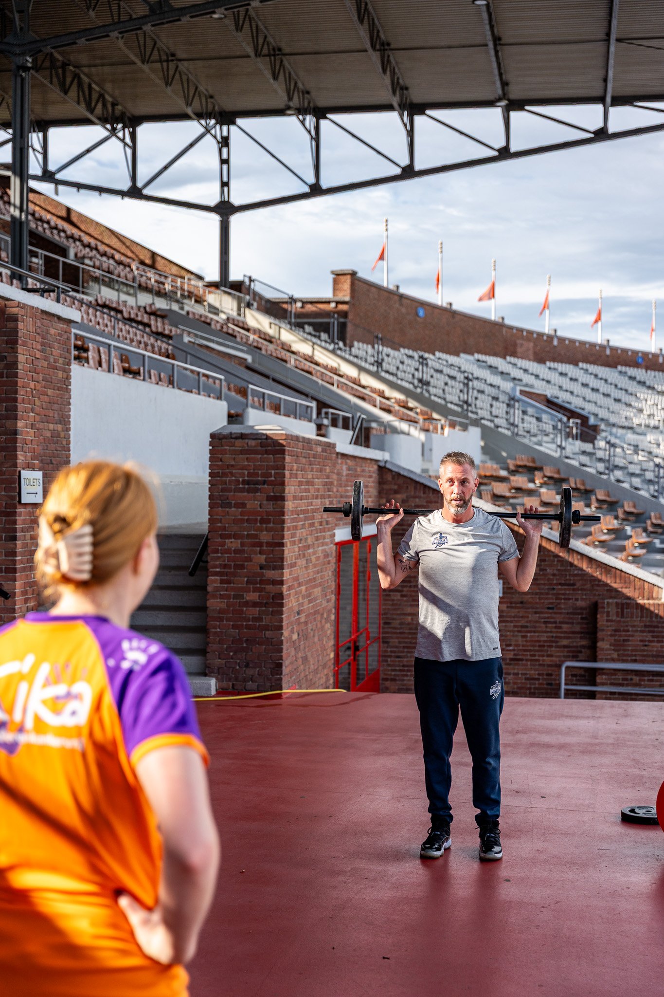 A man is lifting a barbell on his shoulders during a workout at an outdoor stadium, with a young girl watching him. The stadium has empty bleachers and flags in the background.