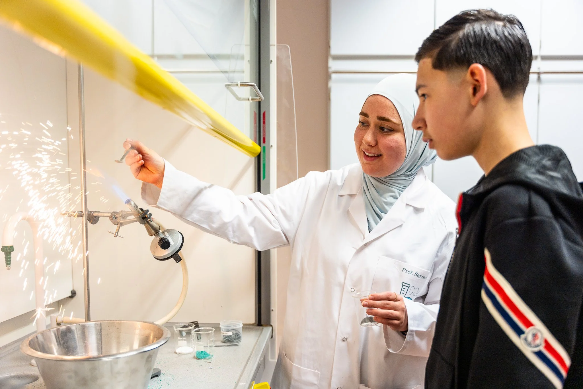 A female scientist in a lab coat and hijab demonstrates a chemical experiment with sparks to a young male student in a black jacket. They are in a laboratory with glassware and equipment on the counter.