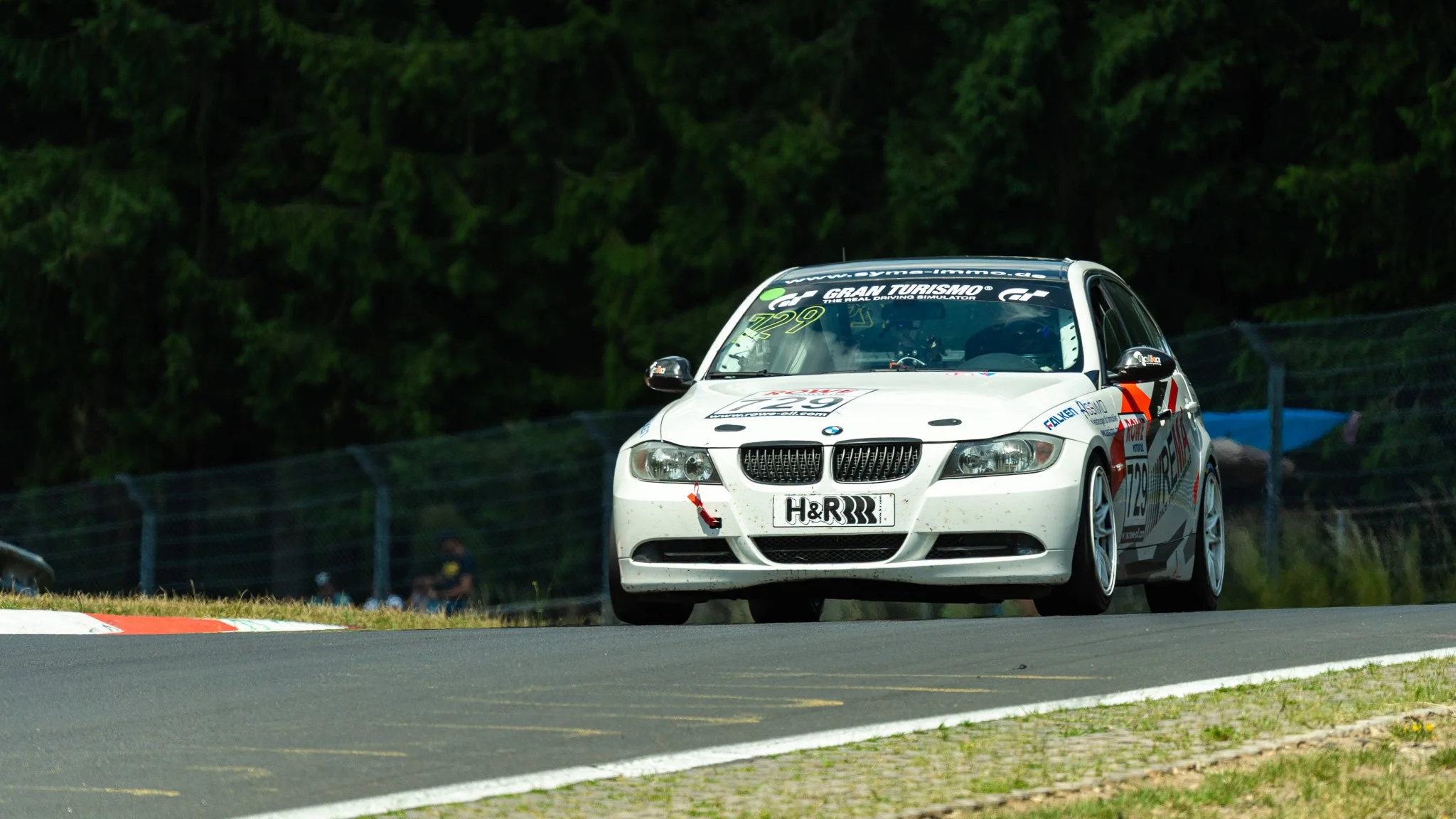 Race car on a track during a race, with a background of trees and a fence, BMW model, with various sponsor decals.