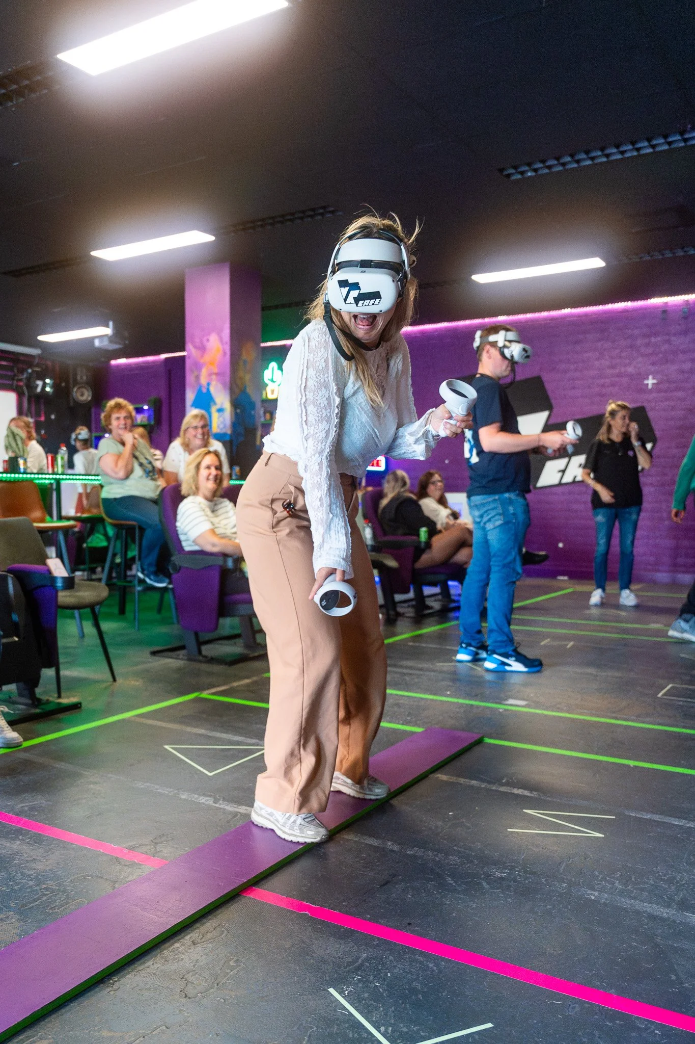People wearing virtual reality headsets and holding controllers in a gaming arcade, with onlookers watching and sitting in chairs.