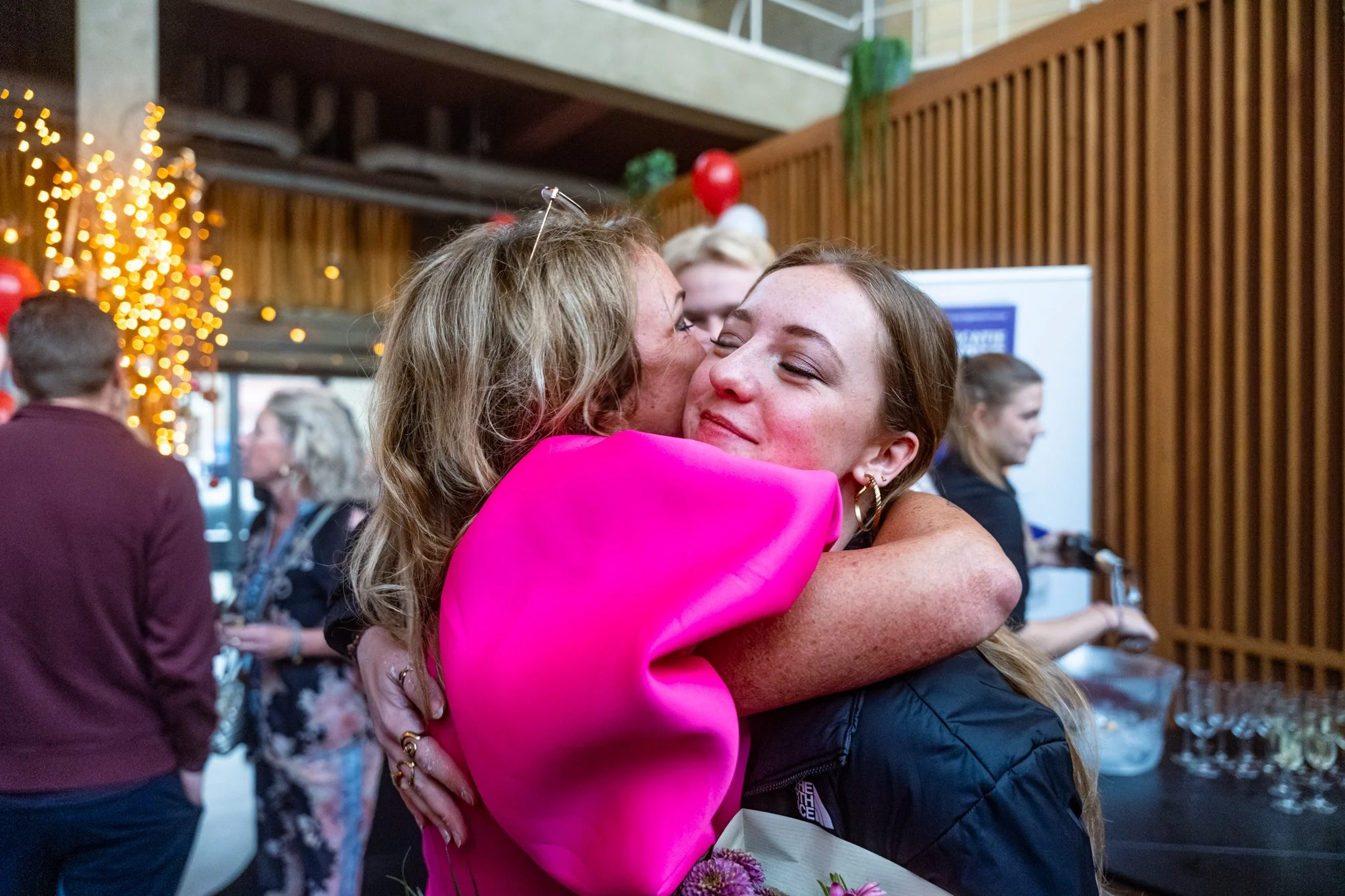 Twee vrouwen omhelzen elkaar in een feestelijke omgeving met kerstversieringen en andere mensen op de achtergrond.