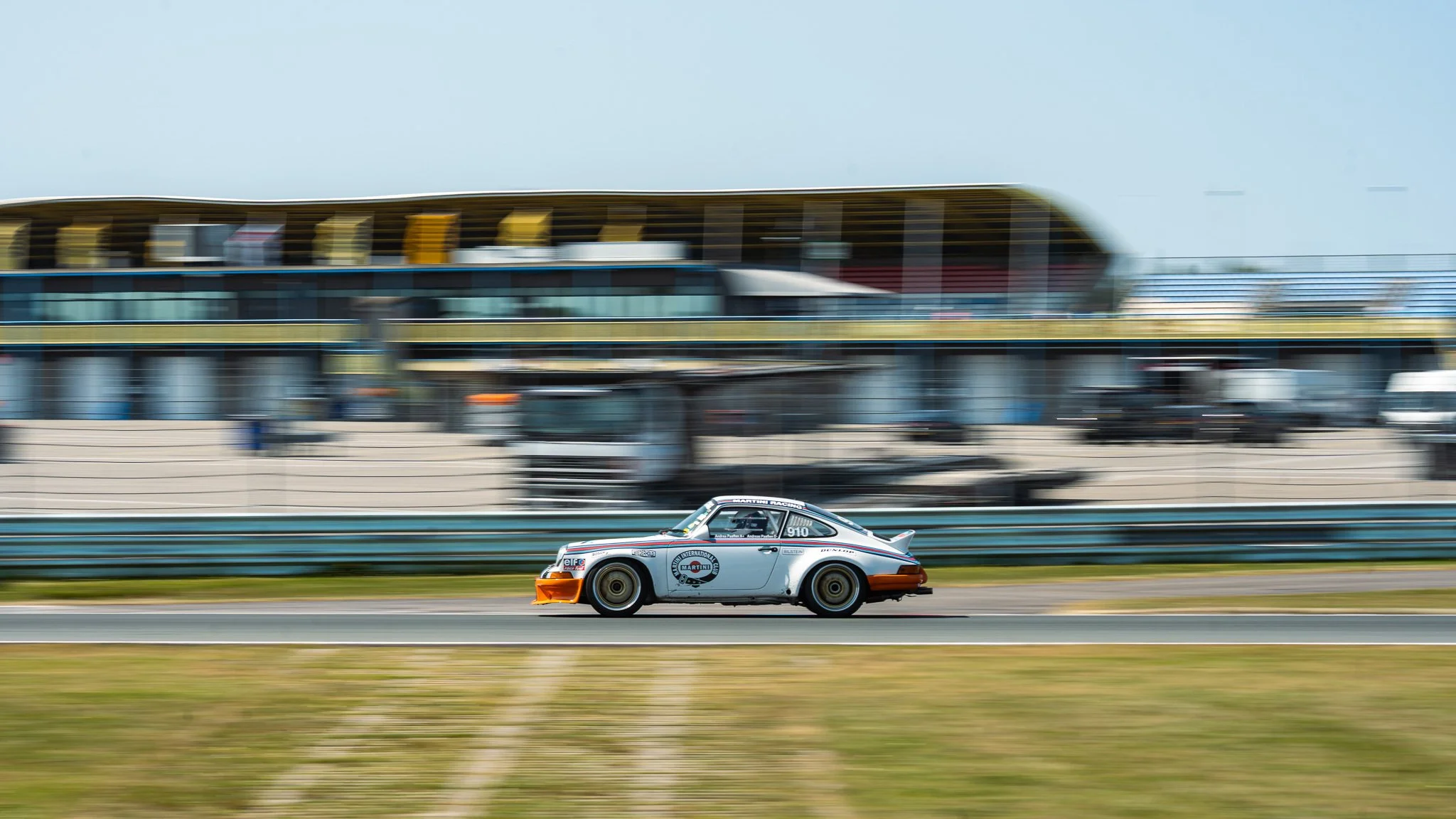 A vintage race car on a track in motion, with a blurred background of a race facility or grandstand, and a blue sky overhead.