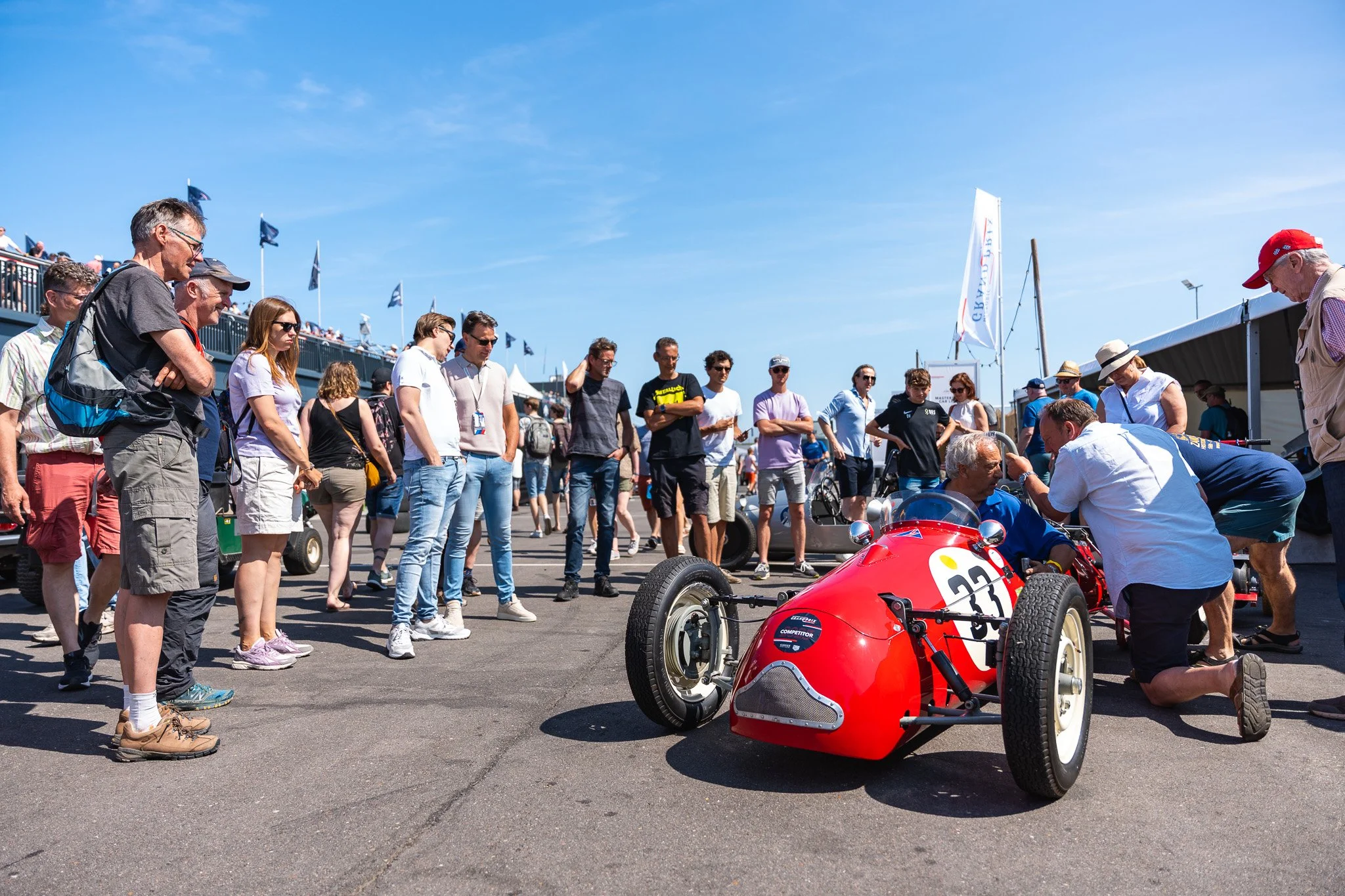 Gathering of people around a vintage red race car at an outdoor event, with some individuals inspecting the car and others watching, under a bright blue sky.