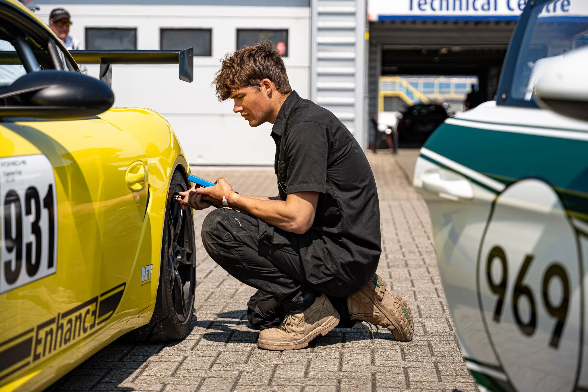 Man knielt op de grond en voert onderhoud uit aan een gele raceauto in een pitbaan, terwijl een vrouw in de achtergrond toekijkt.