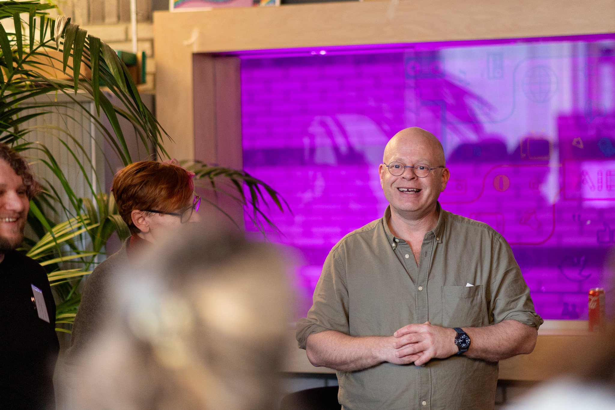 A man with glasses and a beige shirt smiling and clasping his hands together in front of a purple-lit background, surrounded by other people and green plants.