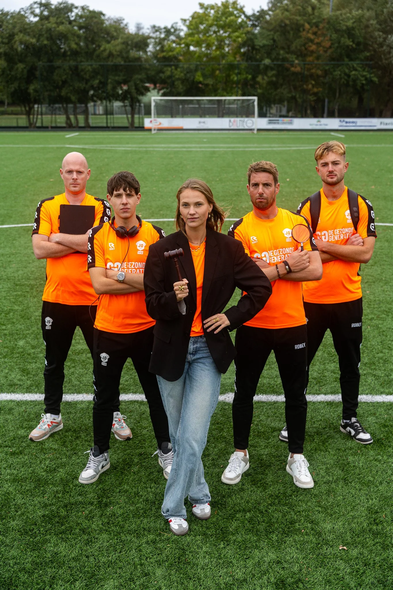 A group of four men and one woman standing on a soccer field, with the woman in front holding a gavel. The men are wearing orange jerseys with black and white details, and the woman is dressed in a black blazer, t-shirt, and jeans. The setting appear