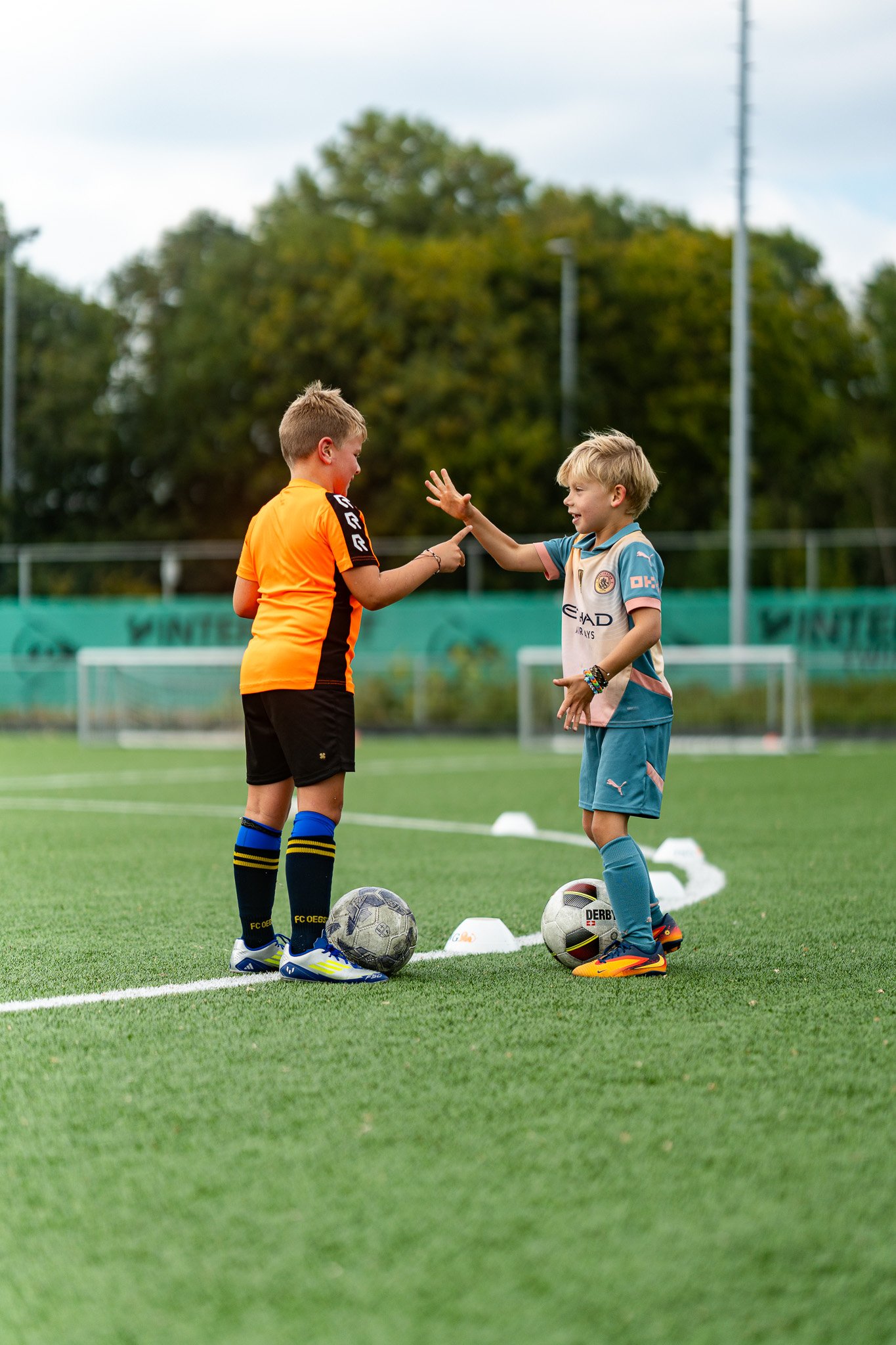 Two young boys in soccer uniforms exchanging high fives on a soccer field, with soccer balls at their feet and cones in the background.