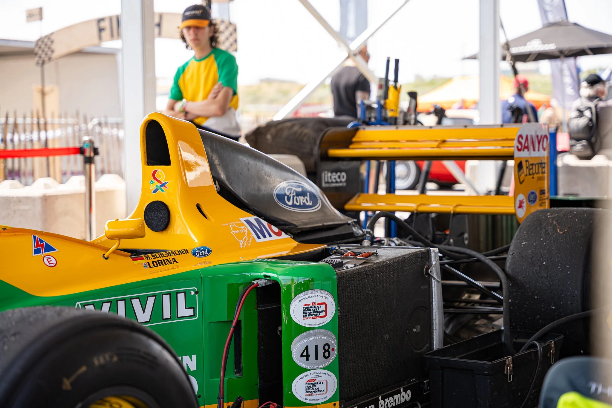 Close-up van een geel en groen raceauto met sponsorstickers en een nummer 418, klaar voor een race, op een pitstraat of paddock, met mensen en tenten op de achtergrond.