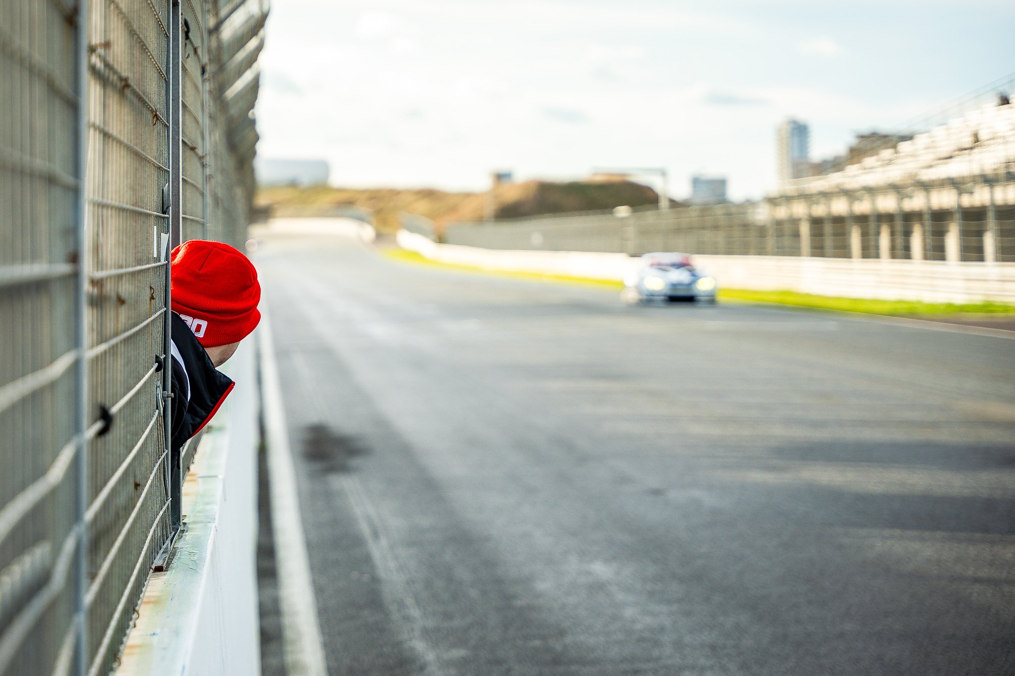 Person wearing a red hat and black jacket leaning on a metal fence along a race track, with a blurred car driving on the track in the background.