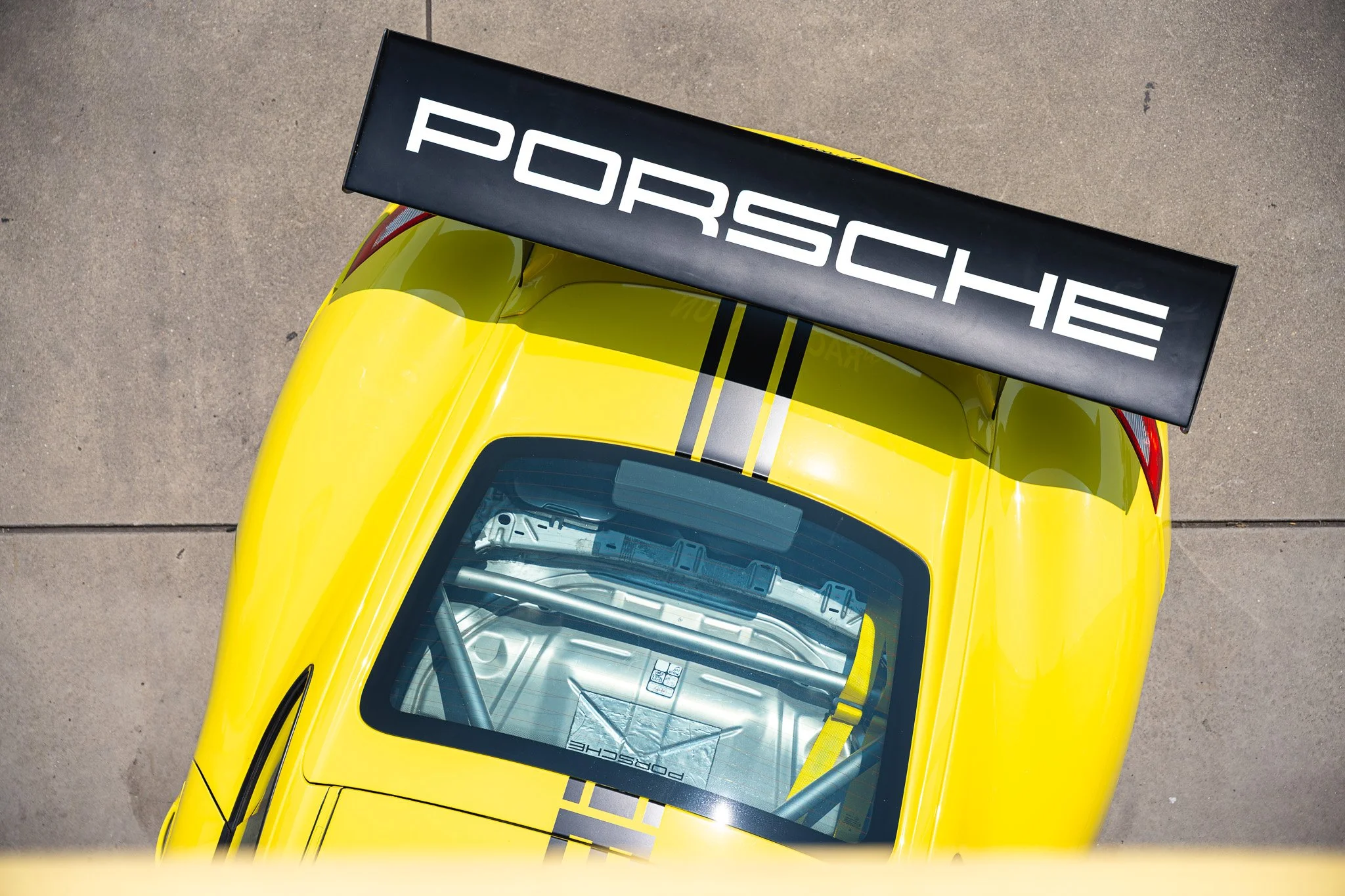 Een geel Porsche raceauto met een grote zwarte achtervleugel waarop de merknaam 'Porsche' staat, gezien vanaf boven op een betonnen surface.
