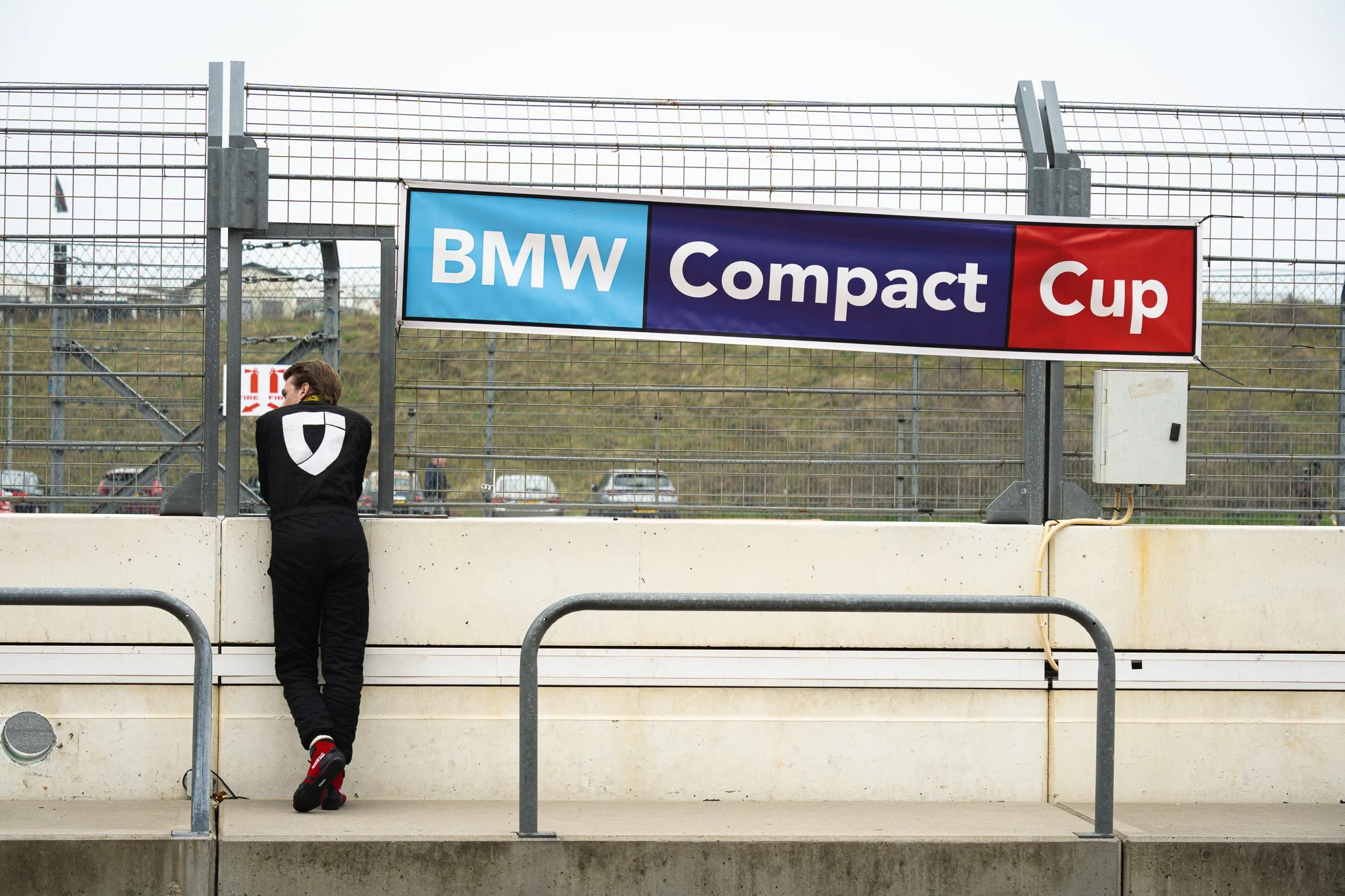 Een persoon in een zwart pak met rode schoenen staat op een trapje bij een hek met een banner erachter die zegt 'BMW Compact Cup'.